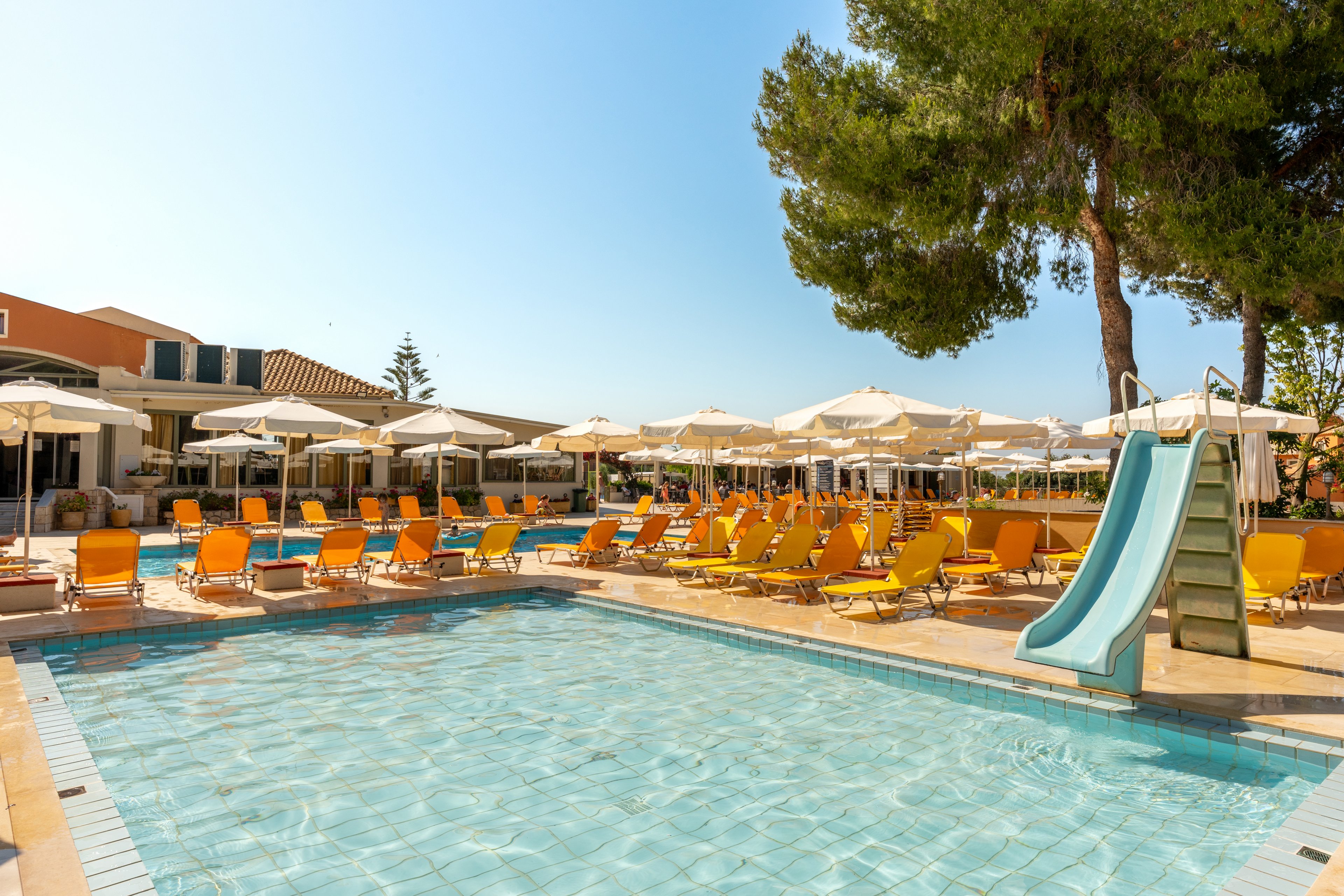 An outdoor resort swimming pool is shown under a clear blue sky, surrounded by yellow sun loungers and white umbrellas. A light blue water slide descends into the shallow end of the pool near a large tree. In the background, low-rise buildings with terraces and additional seating create a relaxed holiday atmosphere. The bright sunlight and warm colors suggest a cheerful, family-friendly vacation setting.