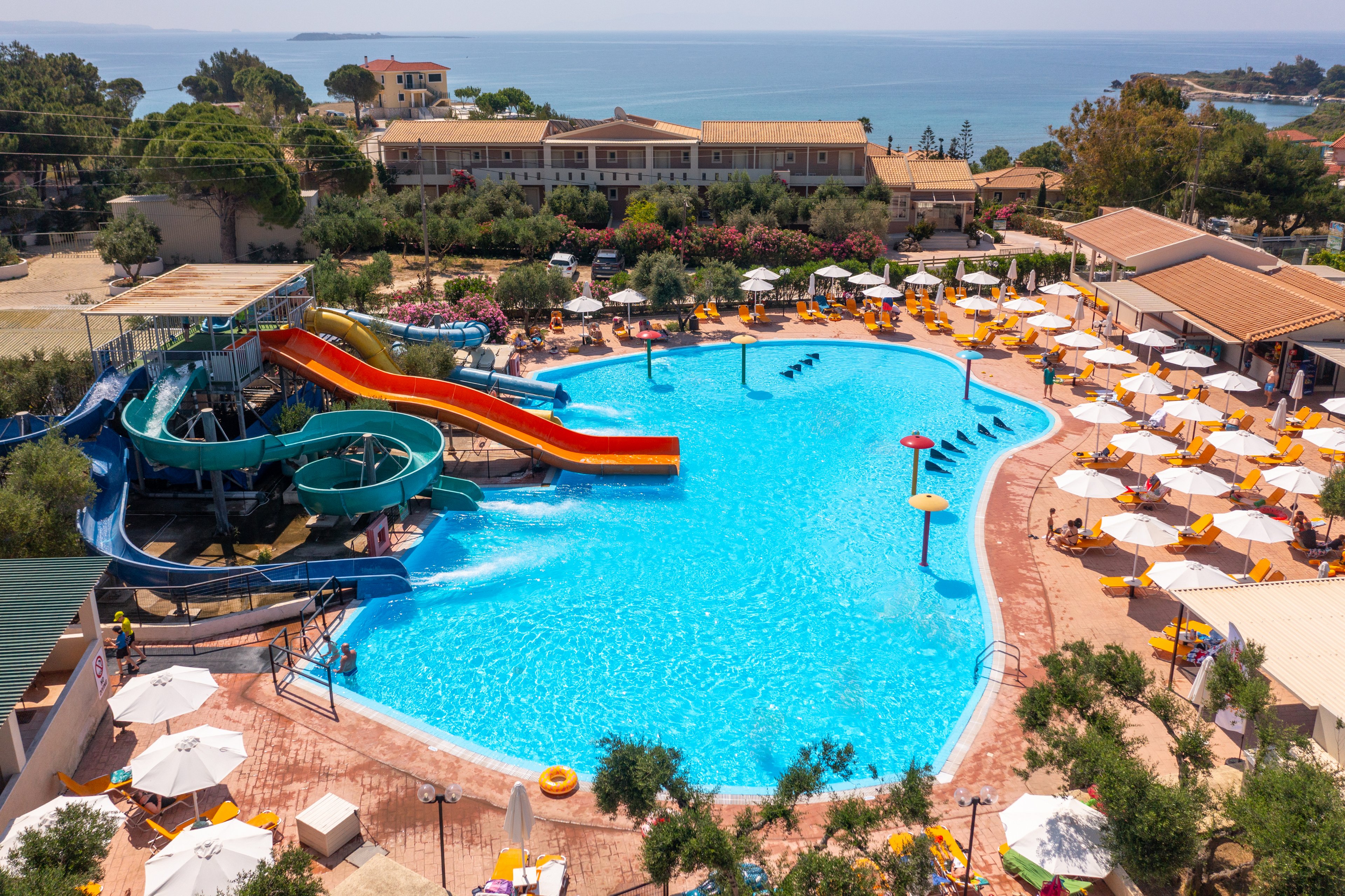 A large outdoor swimming pool is surrounded by sun loungers and umbrellas at a seaside resort. Several bright waterslides curve into the pool, adding a playful attraction for guests. People relax on chairs and enjoy the sunny weather, while the coastline and sea are visible in the background. Trees, buildings, and landscaped paths frame the vibrant holiday setting.