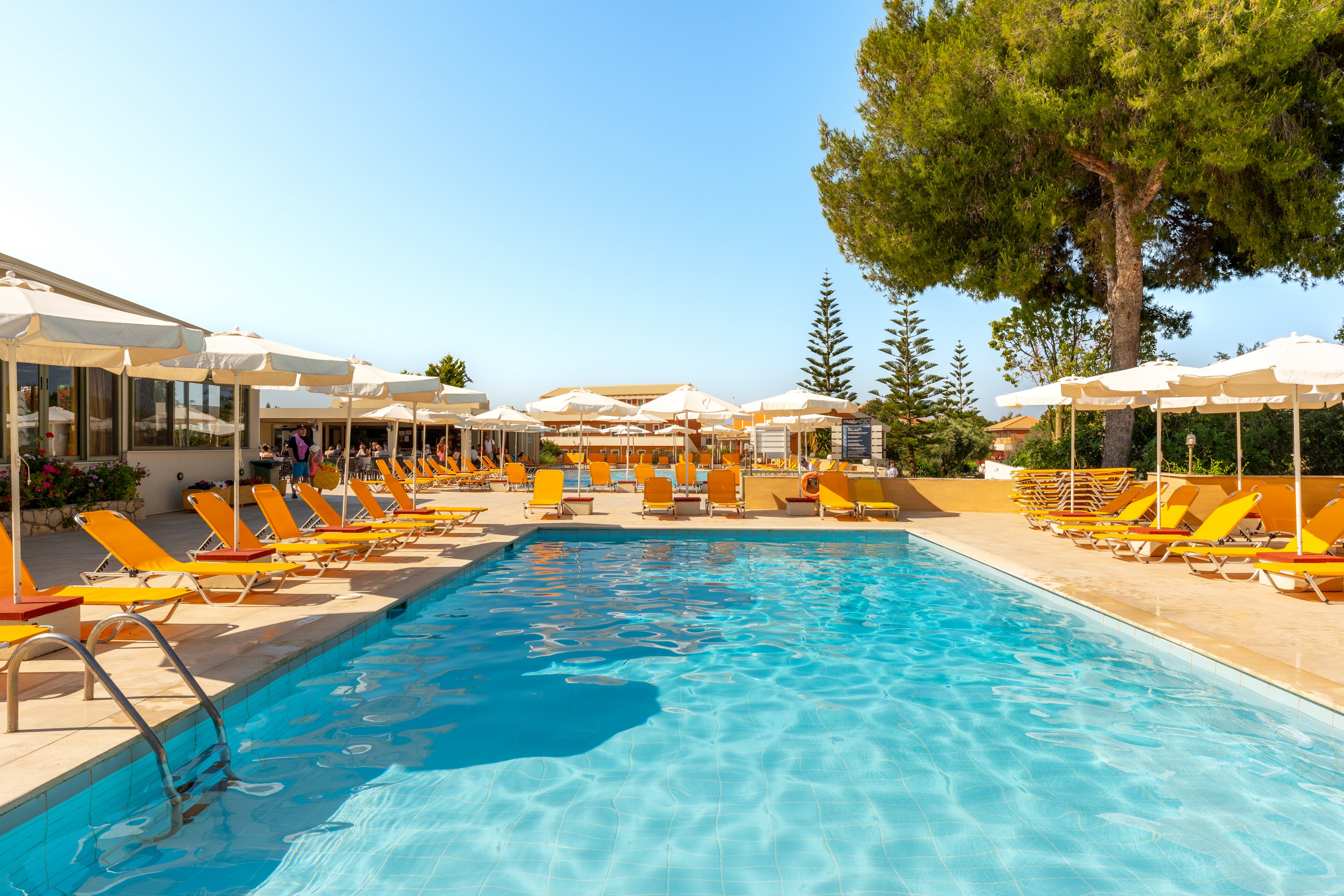 An outdoor swimming pool is surrounded by bright yellow sun loungers and white umbrellas at a holiday resort. The clear blue water reflects the sunny sky, creating a vibrant and inviting atmosphere. Low buildings with large windows line one side of the pool, while tall green trees and shrubs frame the background. The scene suggests a relaxing vacation setting with ample seating and shade for guests.
