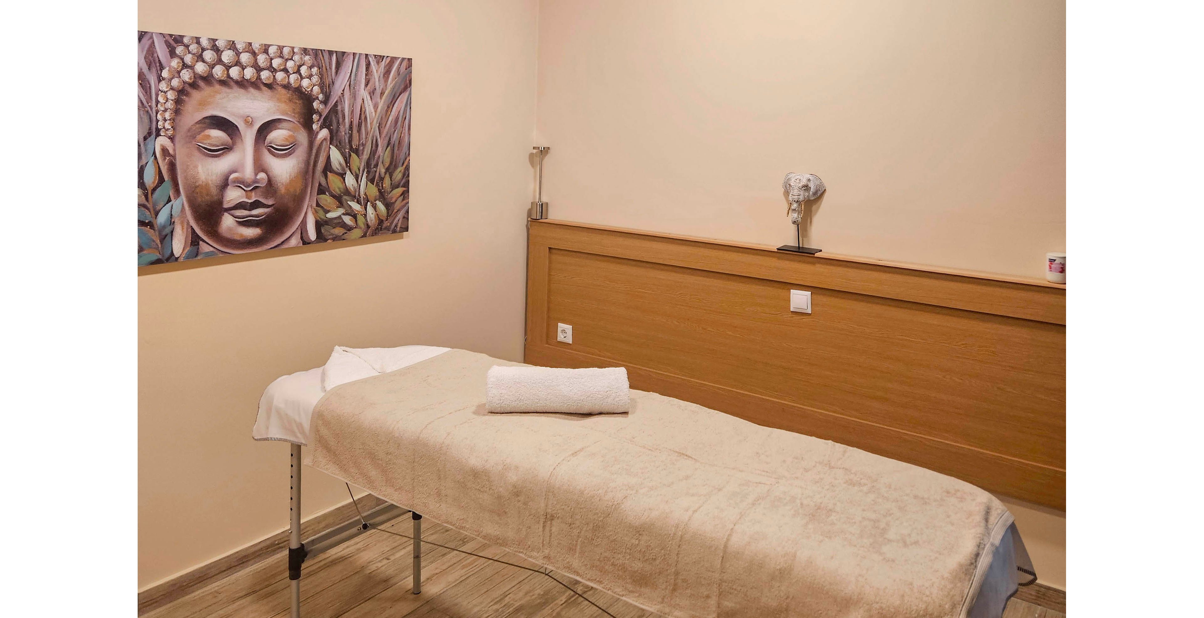 A neatly prepared massage table stands in a small, cozy treatment room. The space features warm beige walls, a wooden accent panel, and a sloped white ceiling with a modern light fixture. A rolled white towel rests at the head of the table, suggesting readiness for a spa or wellness session. Decorative artwork and a small ornamental piece add a calming, minimalist touch to the interior.
