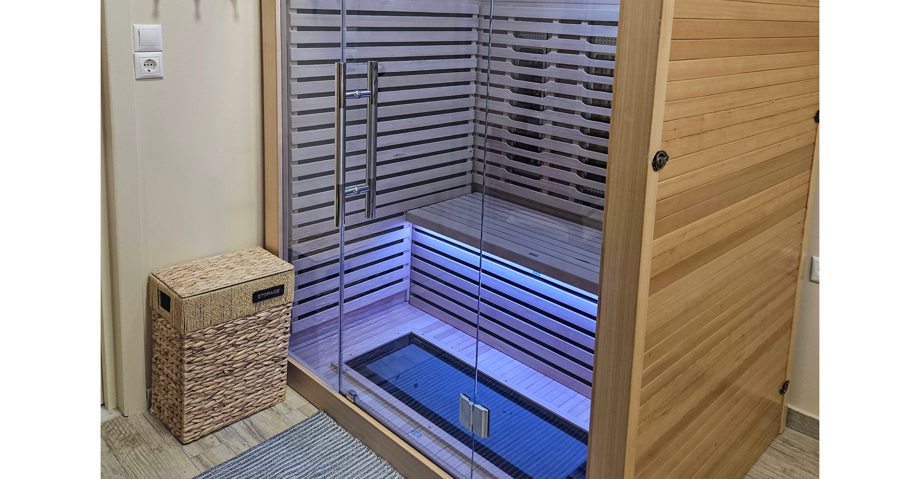 A contemporary wooden infrared sauna cabin stands in a cozy indoor room. The sauna features a large glass front door, interior bench seating, and horizontal slat paneling with integrated lighting. A woven storage basket and a small rug sit on the tiled floor beside the unit, adding to the spa-like atmosphere. The overall scene suggests a private home wellness or relaxation area.