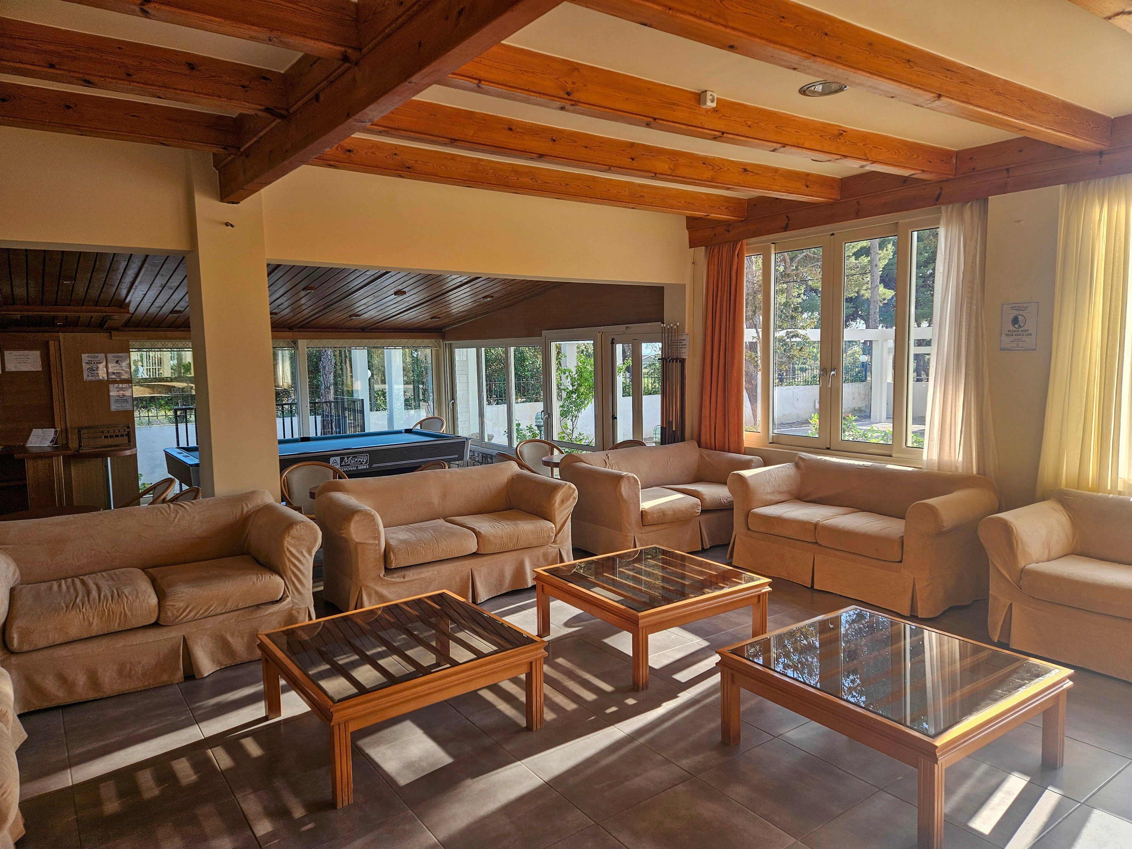 A bright lounge area features multiple beige sofas arranged around glass-top wooden coffee tables. Large windows let in abundant natural light and offer views of trees and an outdoor area. Exposed wooden ceiling beams and tiled flooring create a warm, rustic atmosphere. In the background, a pool table and additional seating suggest a communal or recreational space.