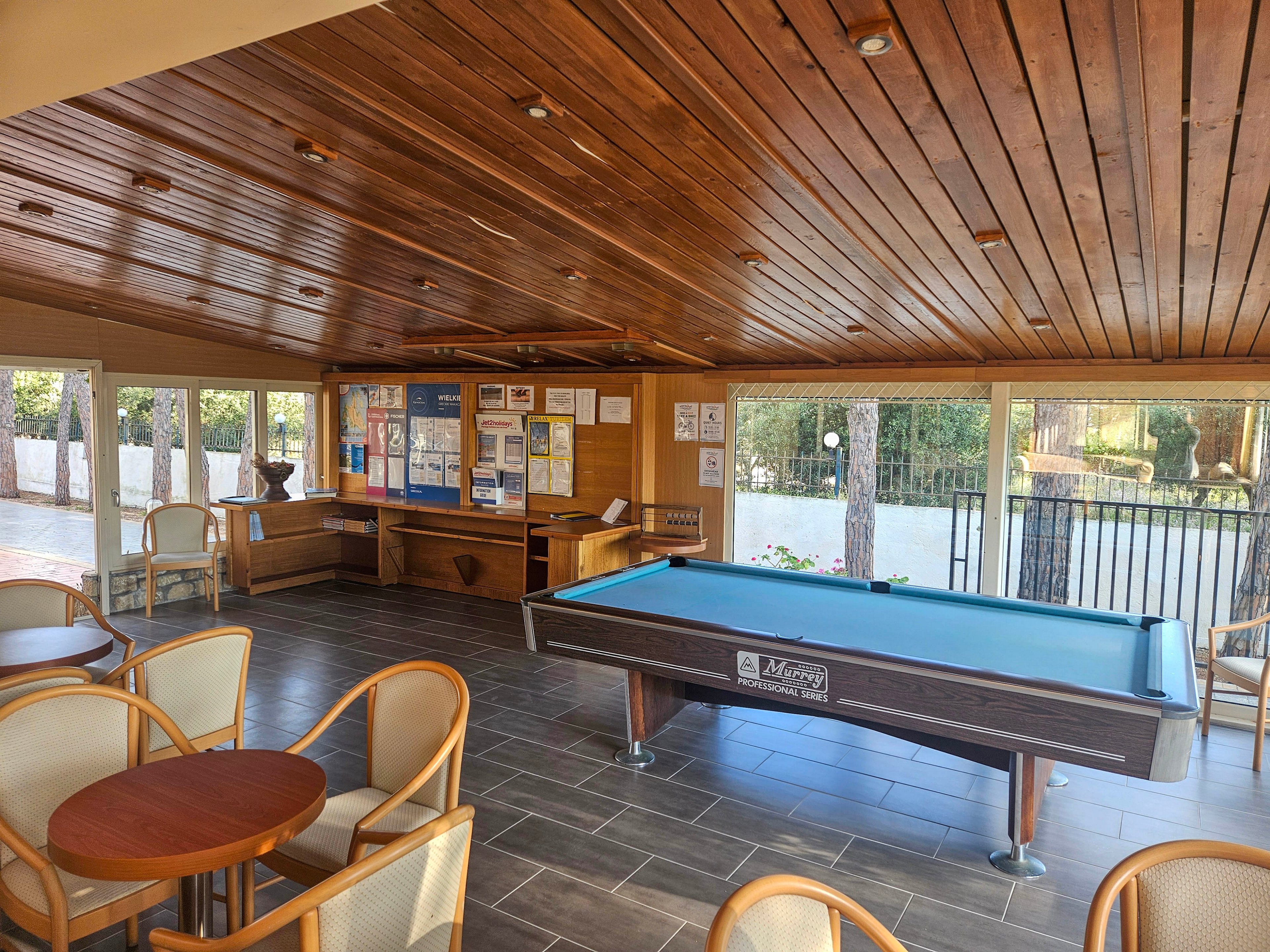 A bright recreation room features a central billiards table with cues resting on its edge. Wooden chairs and round tables are arranged around the space, creating a casual seating area. Large windows and glass doors let in natural light and offer views of trees and an outdoor patio. A wooden ceiling and tiled floor give the room a warm, relaxed atmosphere, while a noticeboard and counter suggest a community or leisure facility.