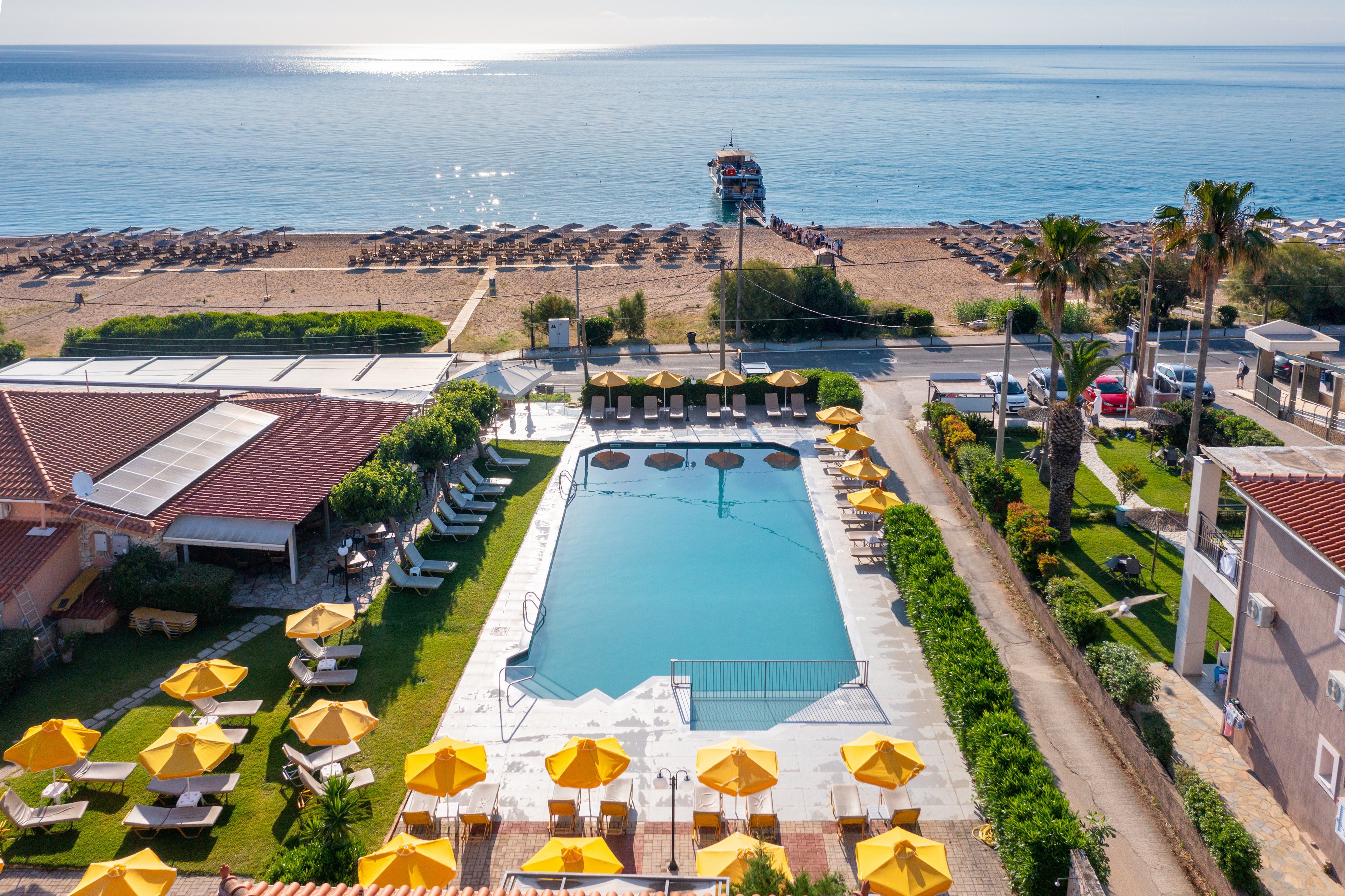 A rectangular swimming pool is surrounded by yellow sun umbrellas and lounge chairs at a beachfront resort. The scene overlooks a sandy beach with rows of sunbeds and the calm sea in the background. Buildings, palm trees, and a coastal road frame the pool area, creating a relaxed holiday atmosphere. Bright sunlight and clear skies emphasize the warm, summery setting.