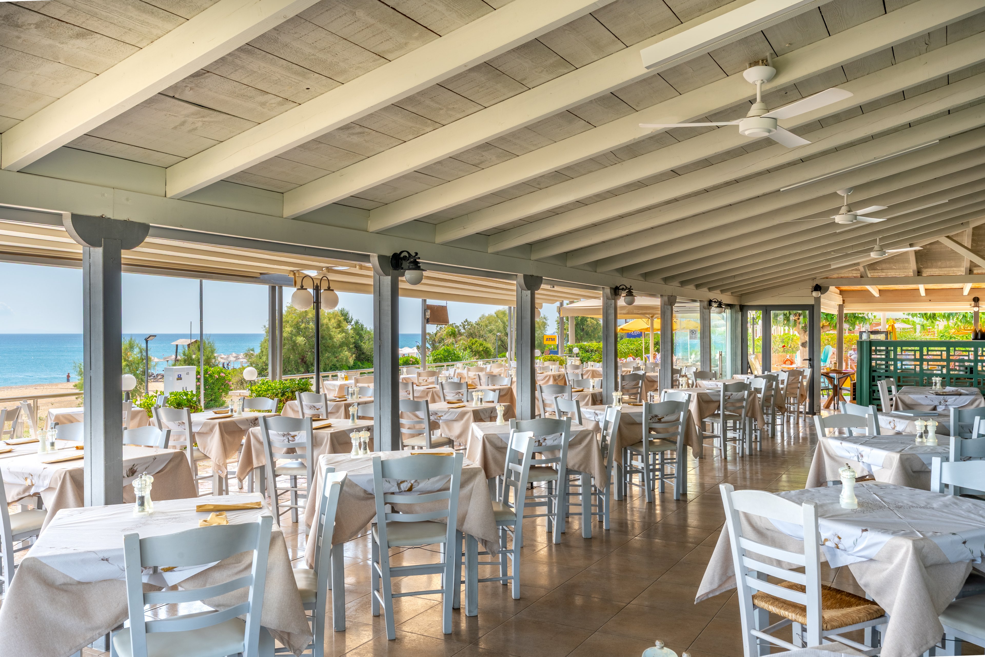 Bright open-air restaurant with rows of white wooden tables and chairs under a sloped ceiling. Beige tablecloths and simple place settings create a casual, coastal dining atmosphere. Large windows and open sides reveal a sandy beach, blue sea, and lush greenery outside. Ceiling fans and natural light enhance the relaxed, summery feel of the space.