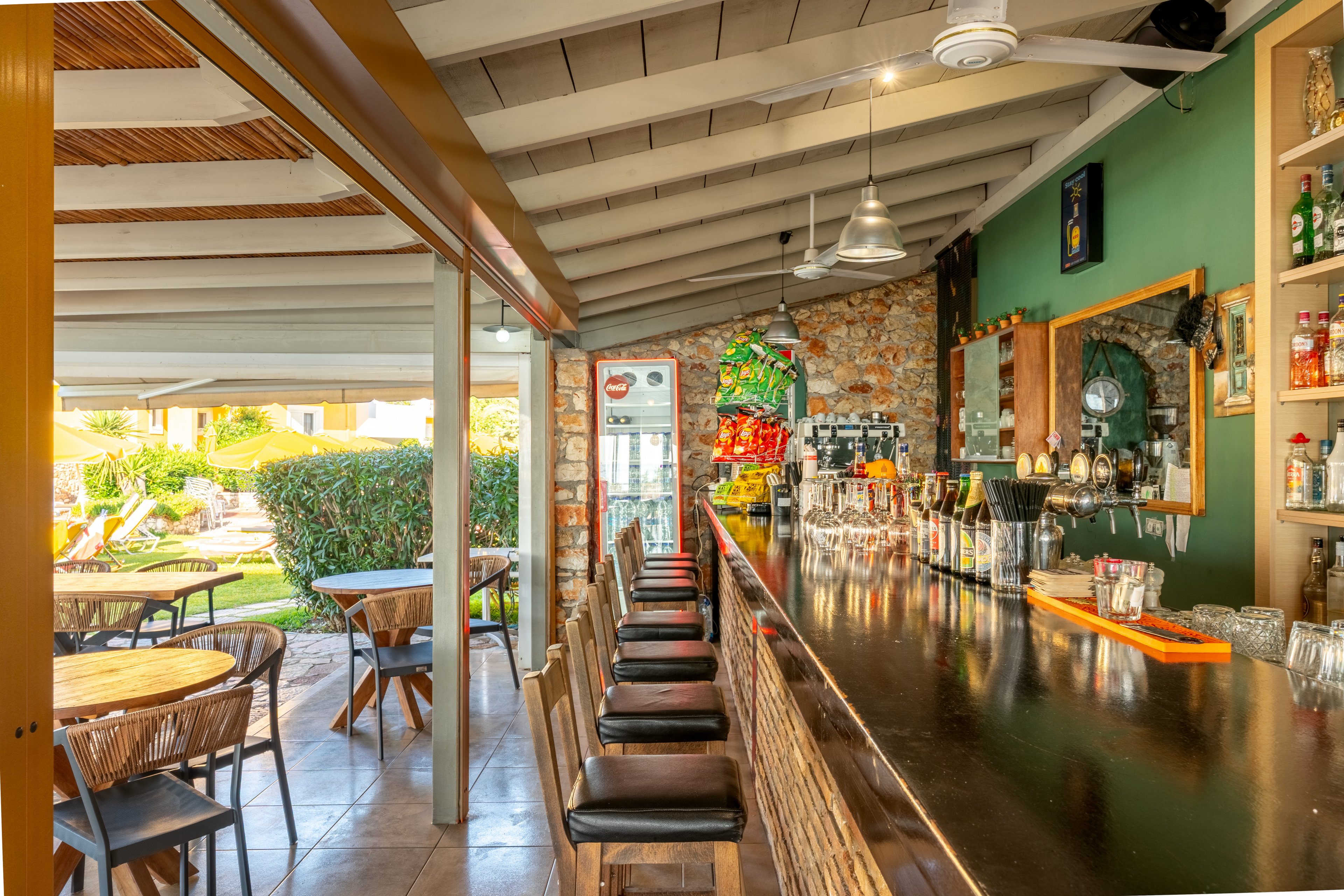 A long polished bar counter with stools stretches through a bright, open-air space. Glassware and bottles are arranged along the bar, with pendant lights hanging from the ceiling and stone walls adding rustic character. To the side, round wooden tables and chairs sit under a covered patio overlooking a green garden. Sunlight fills the area, creating a warm and inviting atmosphere ideal for relaxing drinks.