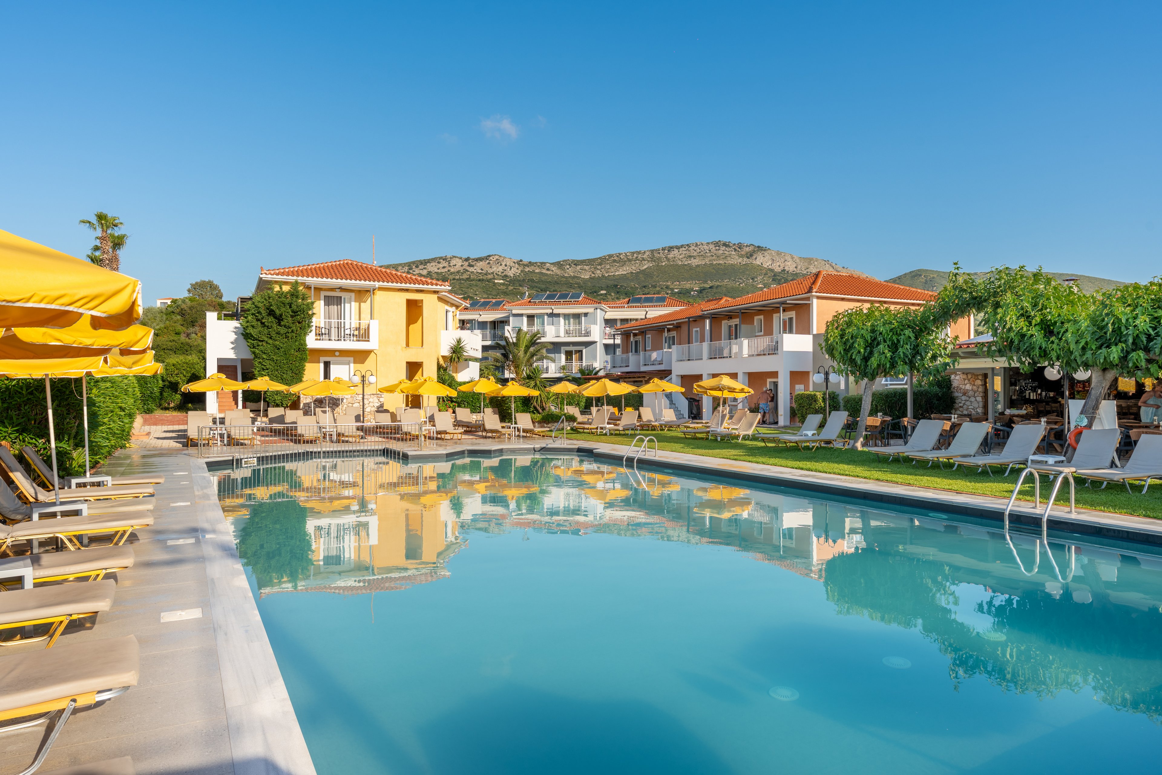 A large outdoor swimming pool reflects surrounding resort buildings under a clear blue sky. Sun loungers with yellow umbrellas line the poolside, creating a relaxed vacation atmosphere. Green trees and shrubs frame the area, with hills and mountains visible in the background. The scene suggests a peaceful holiday setting at a Mediterranean-style hotel.