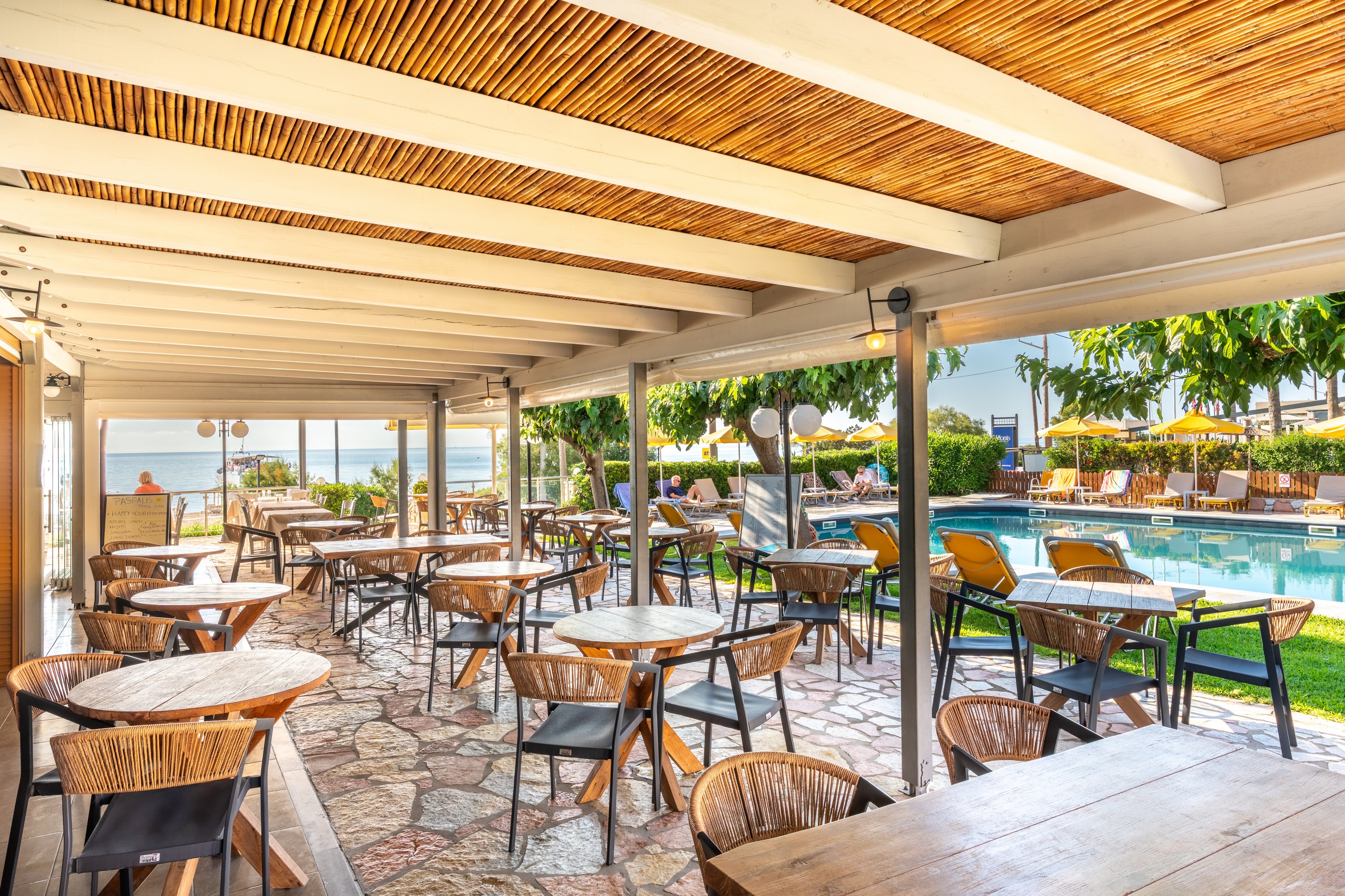 An open-air café with wooden tables and wicker chairs overlooks a bright swimming pool. The covered terrace features a rustic ceiling with wooden slats and stone flooring, creating a relaxed resort atmosphere. Sun loungers and umbrellas line the poolside area, with lush green trees and plants adding to the tropical feel. In the background, the sea and clear blue sky suggest a coastal vacation setting.