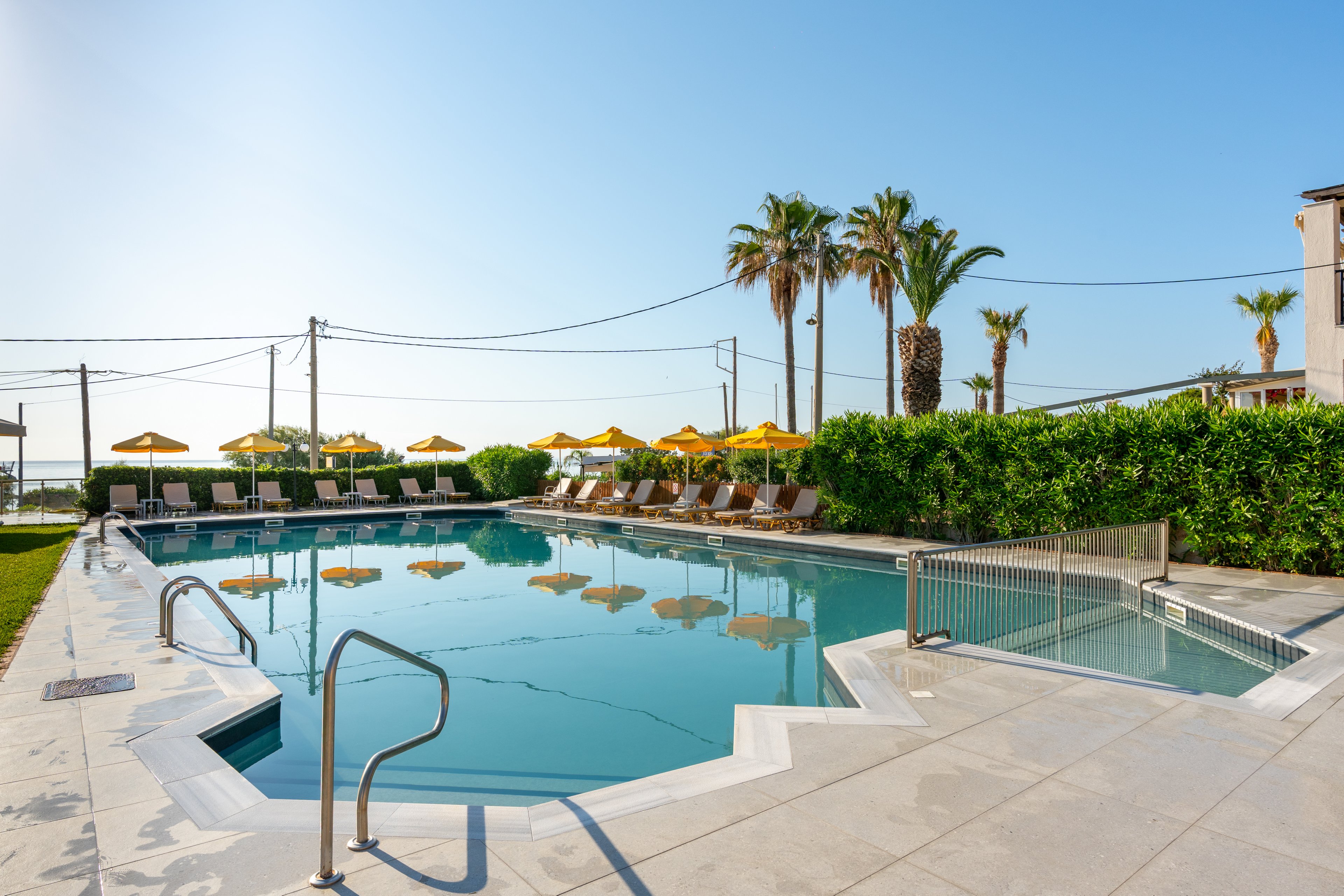 Outdoor resort swimming pool with clear blue water is surrounded by sun loungers and yellow umbrellas. Palm trees rise behind a green hedge, adding a tropical feel to the scene. Metal handrails and a small fenced-off pool area are visible in the foreground. The bright sky and strong sunlight create a relaxed vacation atmosphere near the coast.