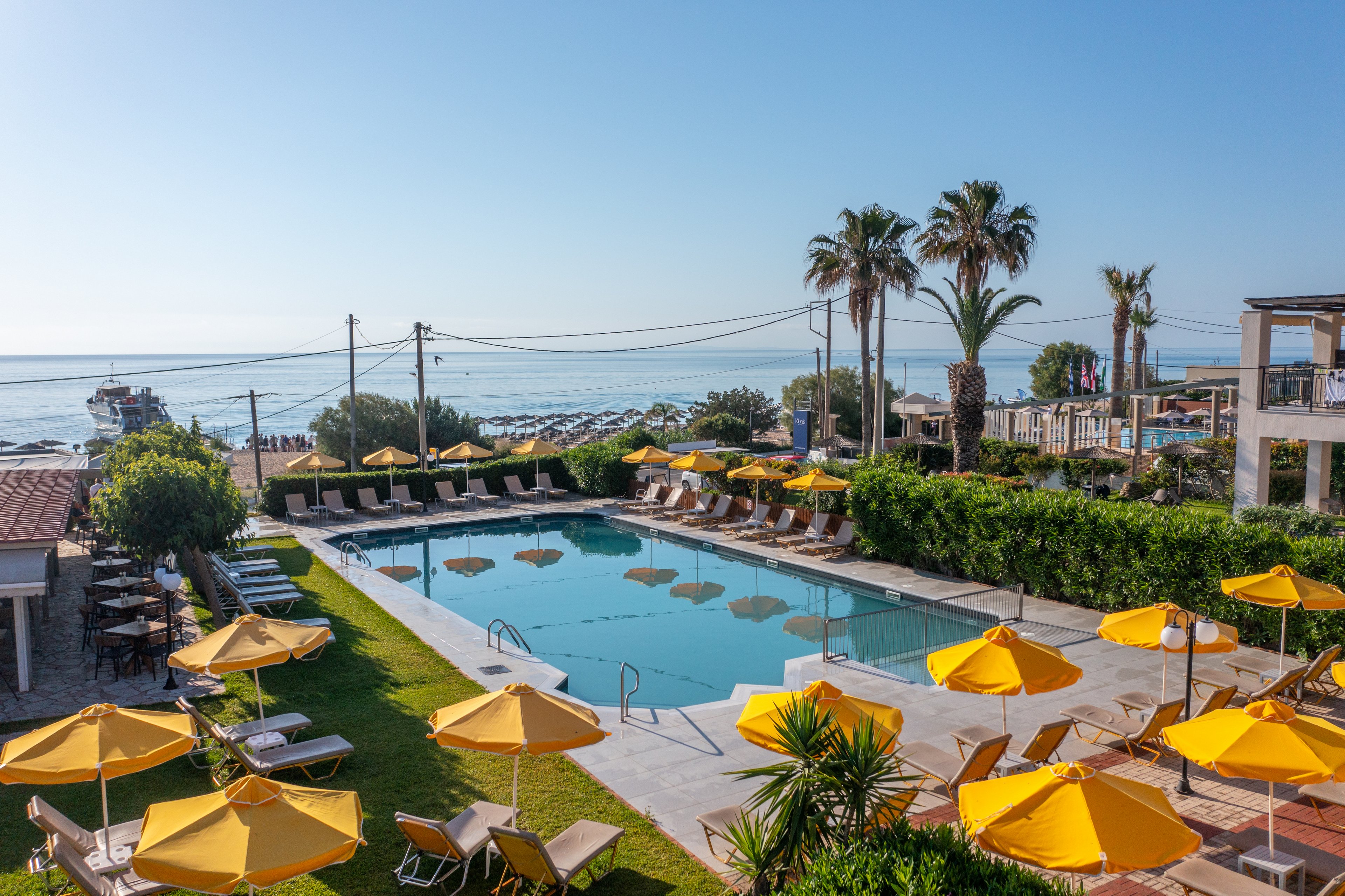An outdoor swimming pool at a seaside resort is surrounded by yellow sun umbrellas and lounge chairs. Palm trees and manicured greenery frame the pool area, creating a relaxed vacation atmosphere. In the background, the beach and calm blue sea are visible under a clear sky. The scene suggests a warm, sunny day ideal for leisure and sunbathing.