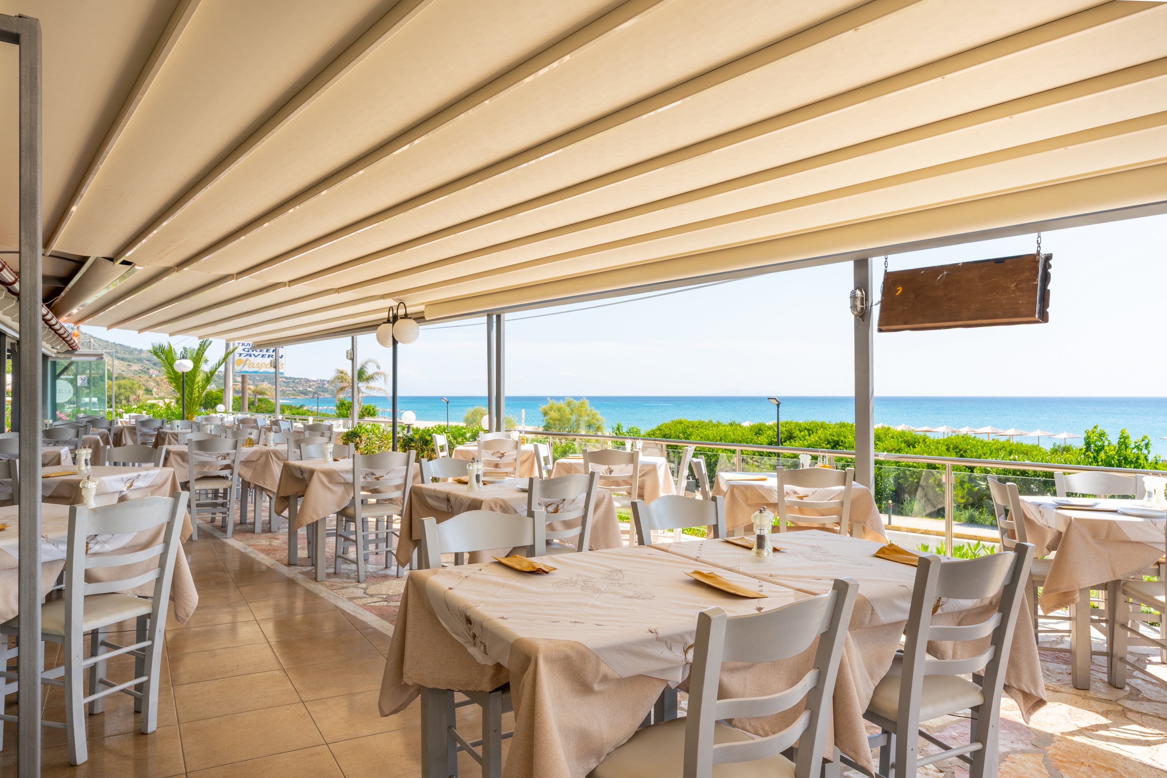 An open-air restaurant terrace features neatly arranged tables covered with beige tablecloths and white chairs. The space is shaded by a large retractable awning, creating a comfortable dining area. In the background, lush greenery and a clear blue sea are visible under a bright sky, suggesting a coastal location. The scene is calm and inviting, with no people present and table settings prepared for future guests.