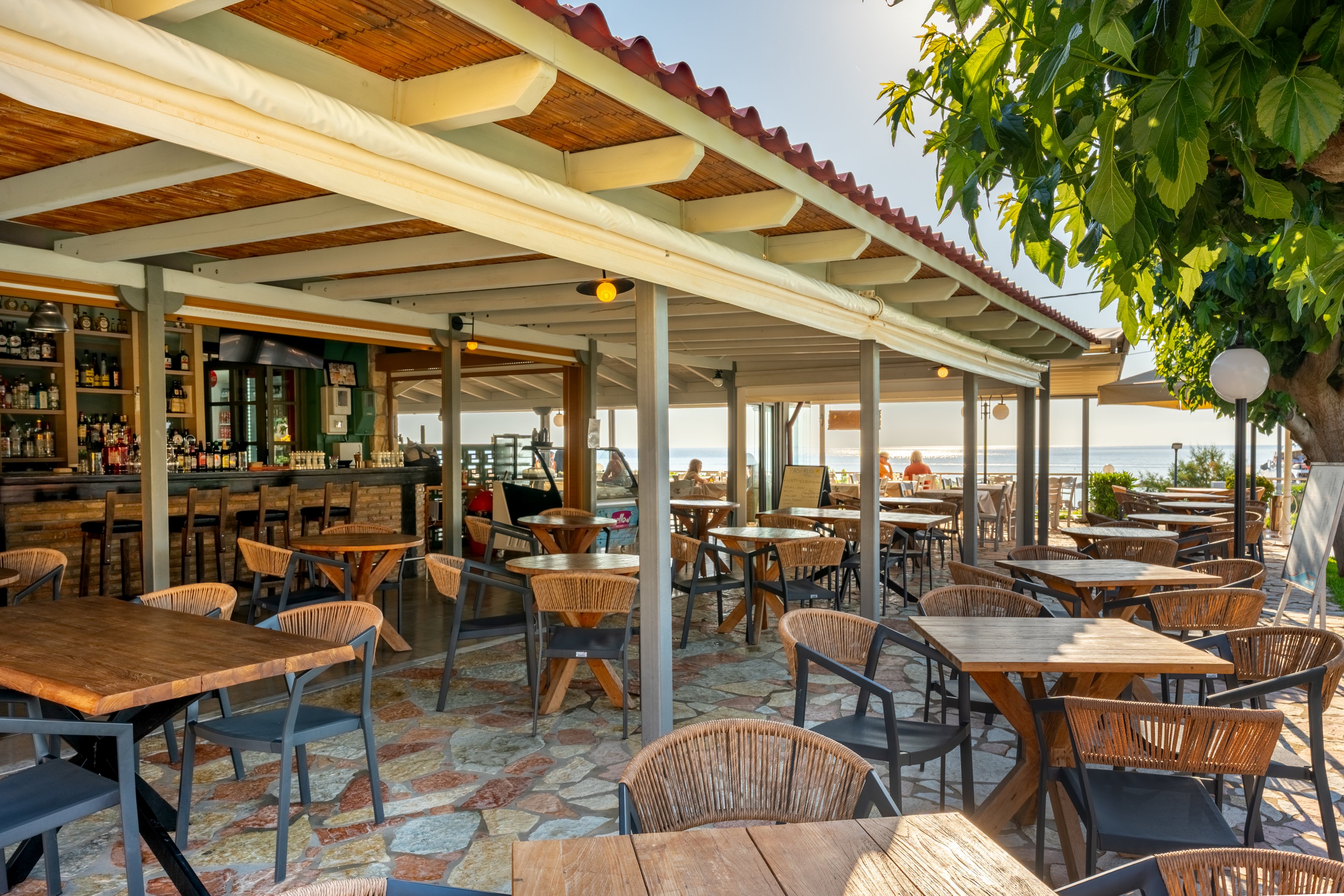 An open-air café with wooden tables and wicker chairs sits under a covered terrace near the sea. The bar area in the background is stocked with bottles and glassware, while the seating area is currently empty. Sun loungers and umbrellas are visible beyond the railing, suggesting a beachfront location. Warm sunlight and natural materials create a relaxed, holiday atmosphere.