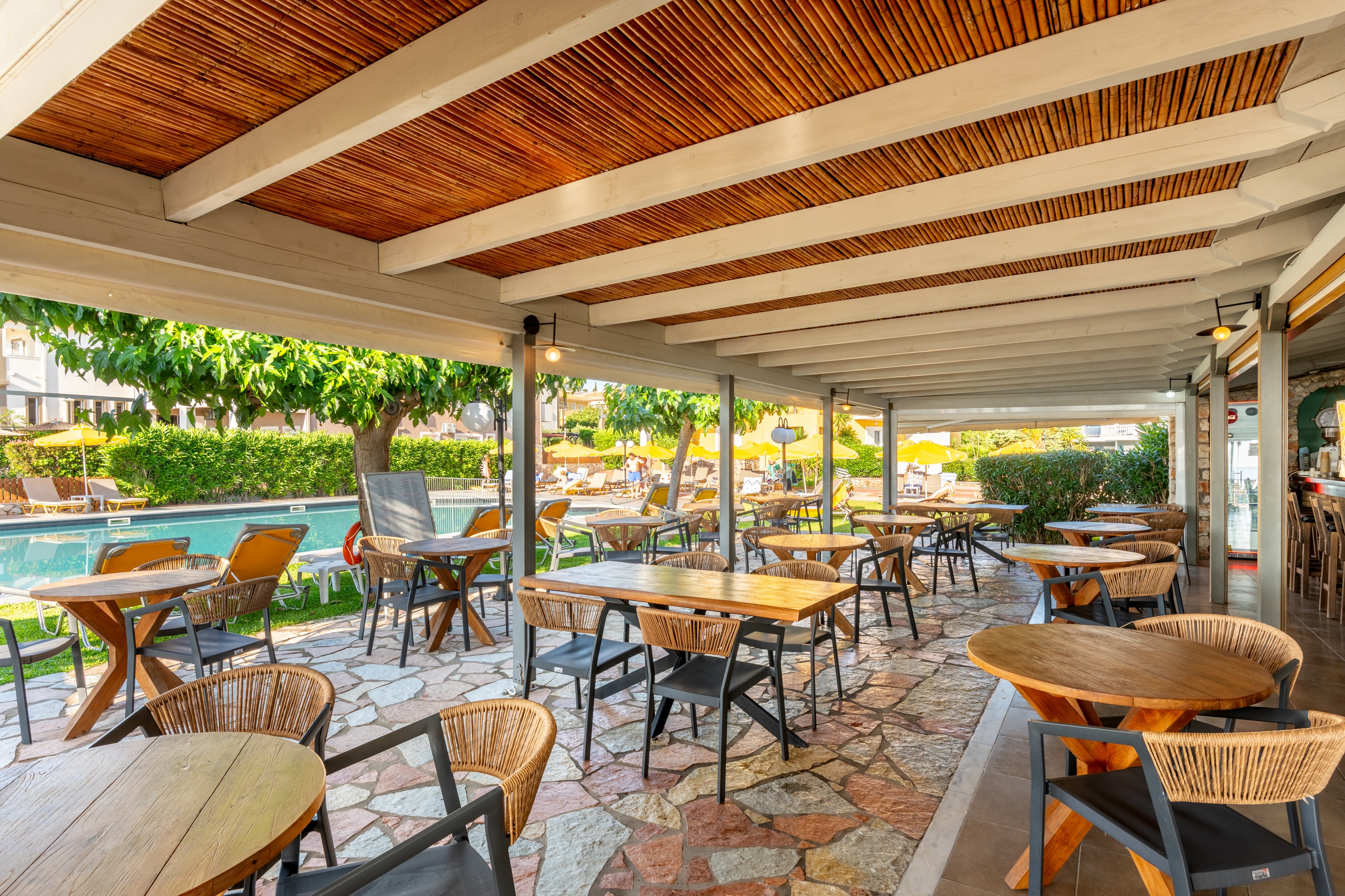 An inviting outdoor café terrace features wooden tables and chairs arranged under a slatted wooden ceiling. The space overlooks a bright poolside area with sun loungers and lush green trees in the background. Warm sunlight enhances the natural wood tones and stone flooring, creating a relaxed vacation atmosphere. The scene suggests a quiet, comfortable spot for dining or enjoying drinks outdoors.