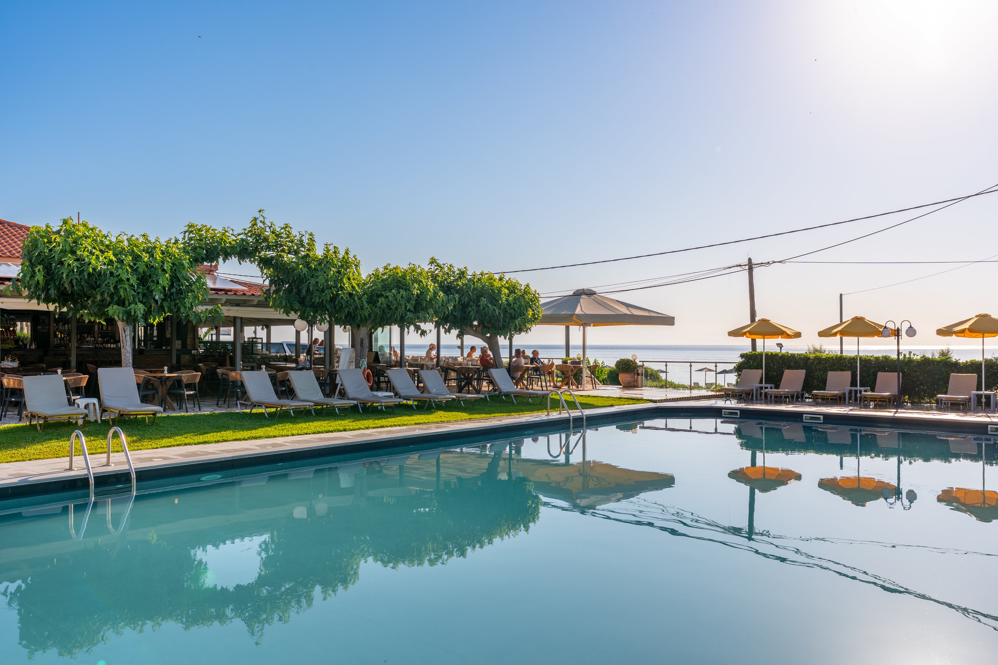 An outdoor swimming pool reflects the clear blue sky at a sunny resort. White lounge chairs and umbrellas line the poolside, offering shaded seating for guests. A covered terrace with tables and greenery sits behind the chairs, suggesting a bar or dining area. In the distance, the sea and horizon are visible, creating a relaxed vacation atmosphere.