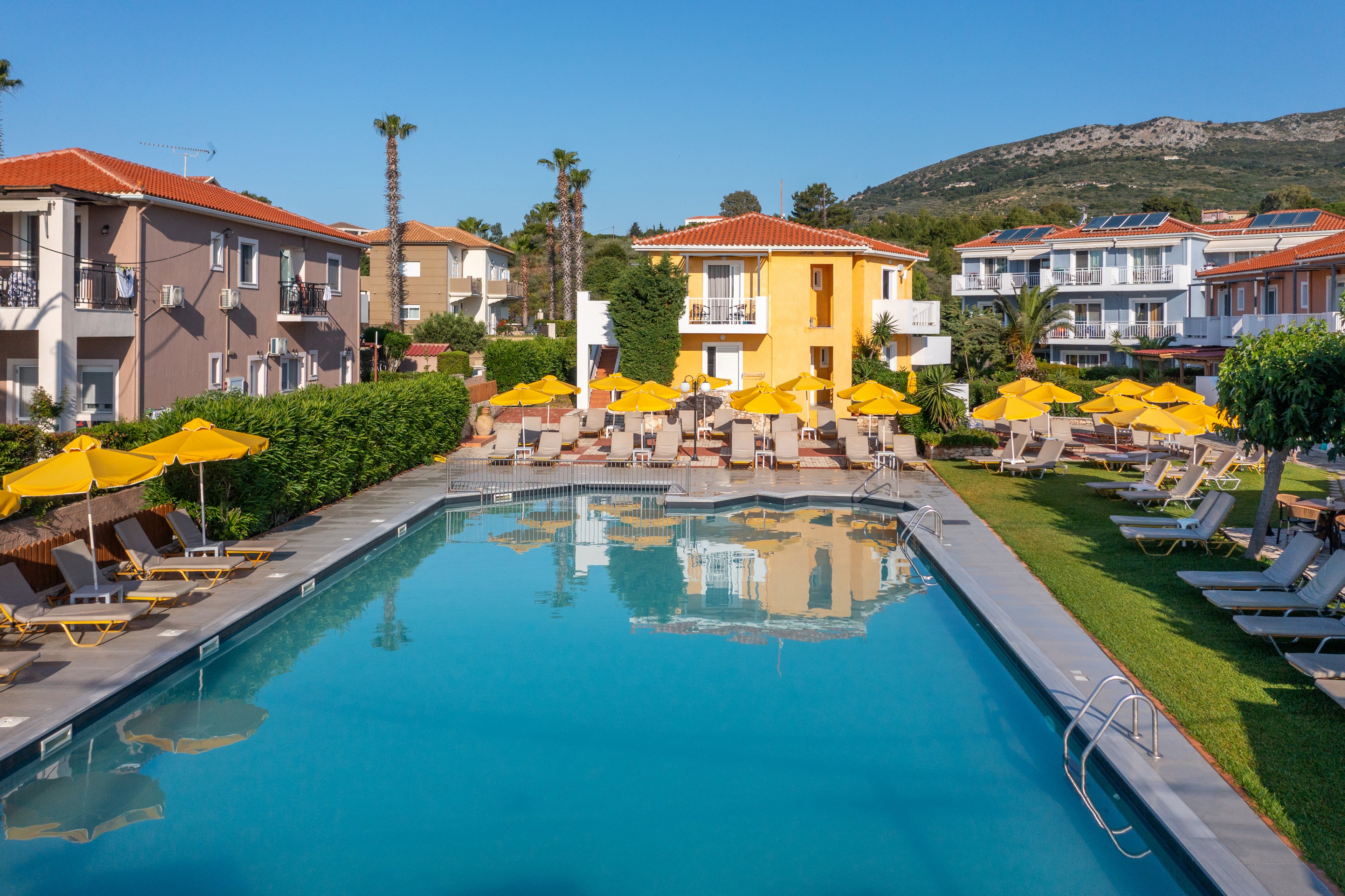 A large outdoor swimming pool is surrounded by sun loungers and bright yellow umbrellas at a holiday resort. Low-rise buildings with red-tiled roofs frame the pool area, with balconies overlooking the water. Green lawns, hedges, and palm trees add a lush feel to the setting, while distant hills rise in the background. The clear blue sky and calm water create a relaxed, inviting vacation atmosphere.
