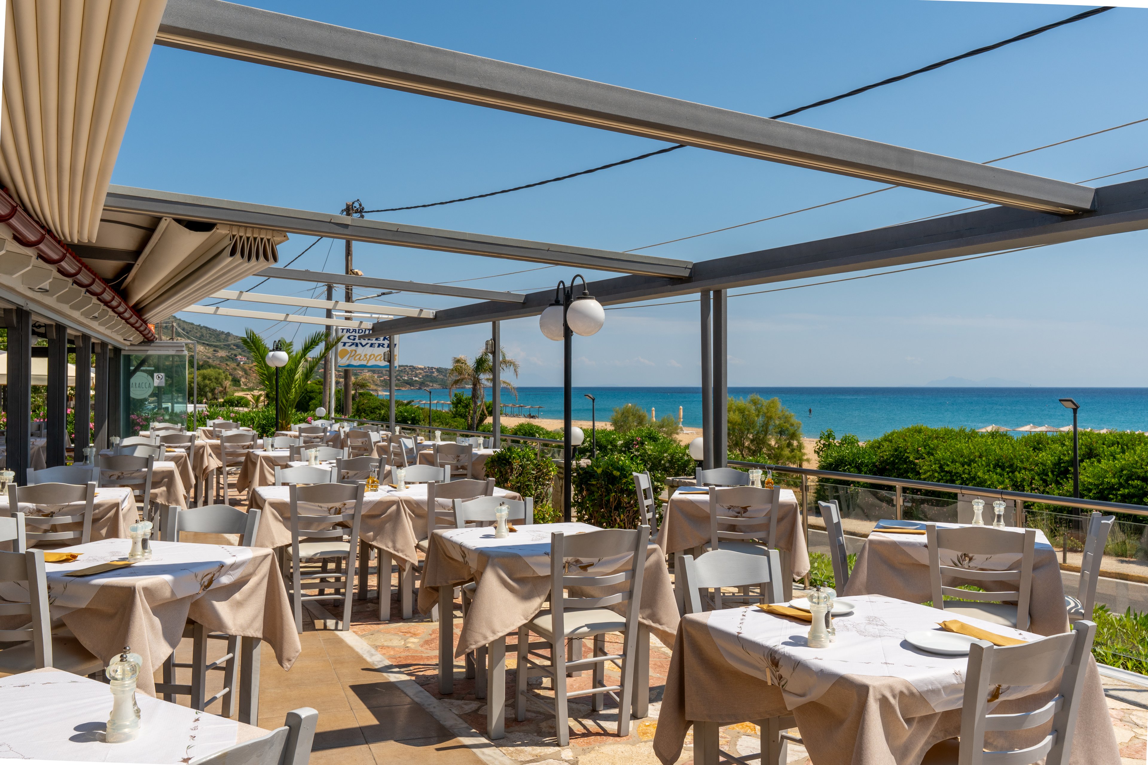 An open-air restaurant terrace is set with neatly arranged tables and chairs overlooking a bright blue sea. Beige tablecloths and white furniture create a light, coastal atmosphere under a partially covered pergola structure. The scene is sunny and calm, with green shrubs and a sandy beach visible beyond the railing. No people are present, emphasizing the tranquil, inviting setting for seaside dining.