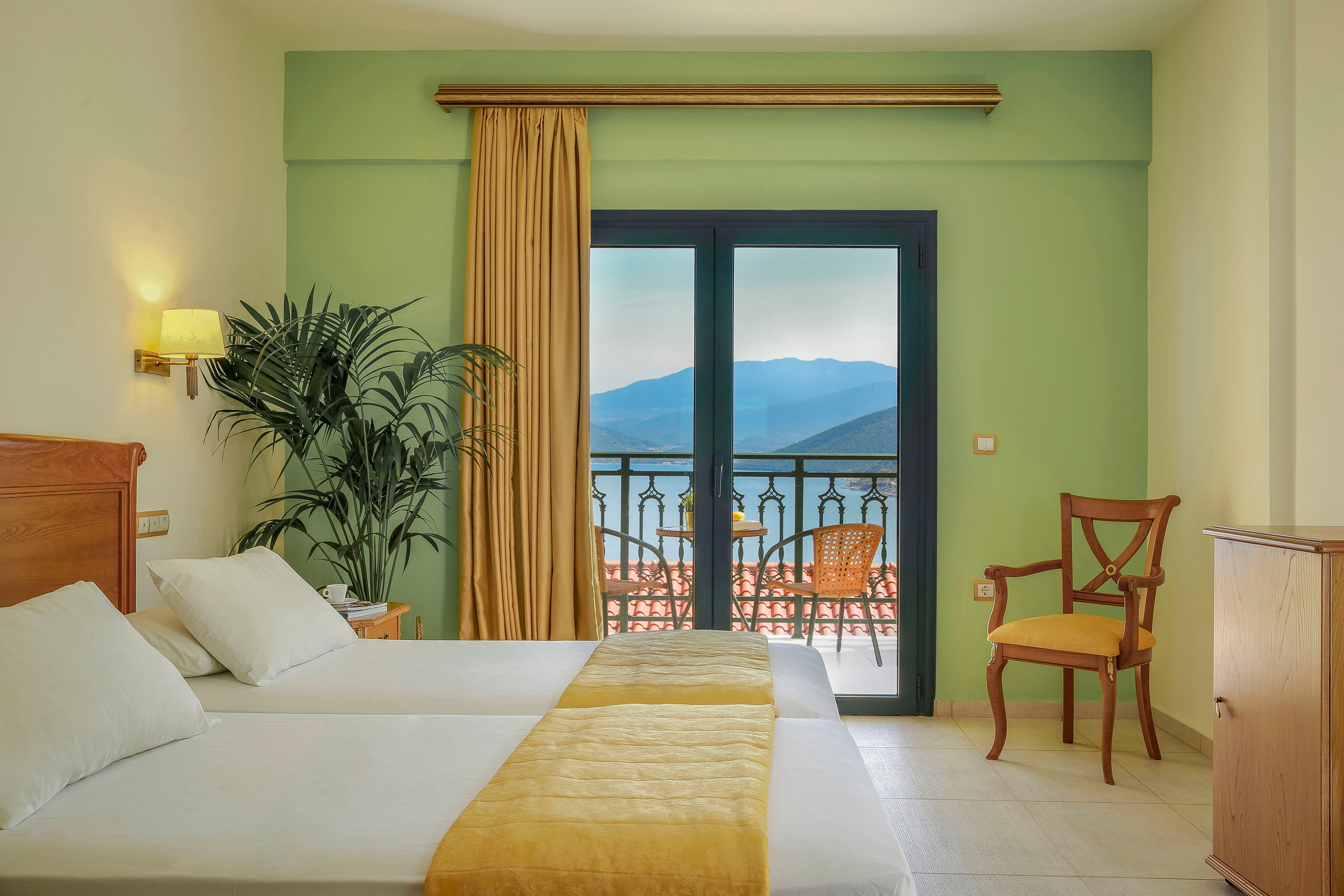 A tidy hotel room features two single beds with white linens and a yellow runner. The space is decorated in soft green and cream tones, with a wooden chair and a potted plant adding warmth. Large glass doors open onto a balcony with outdoor chairs and a railing, overlooking a calm sea and distant mountains. Natural light fills the room, creating a relaxed vacation atmosphere.