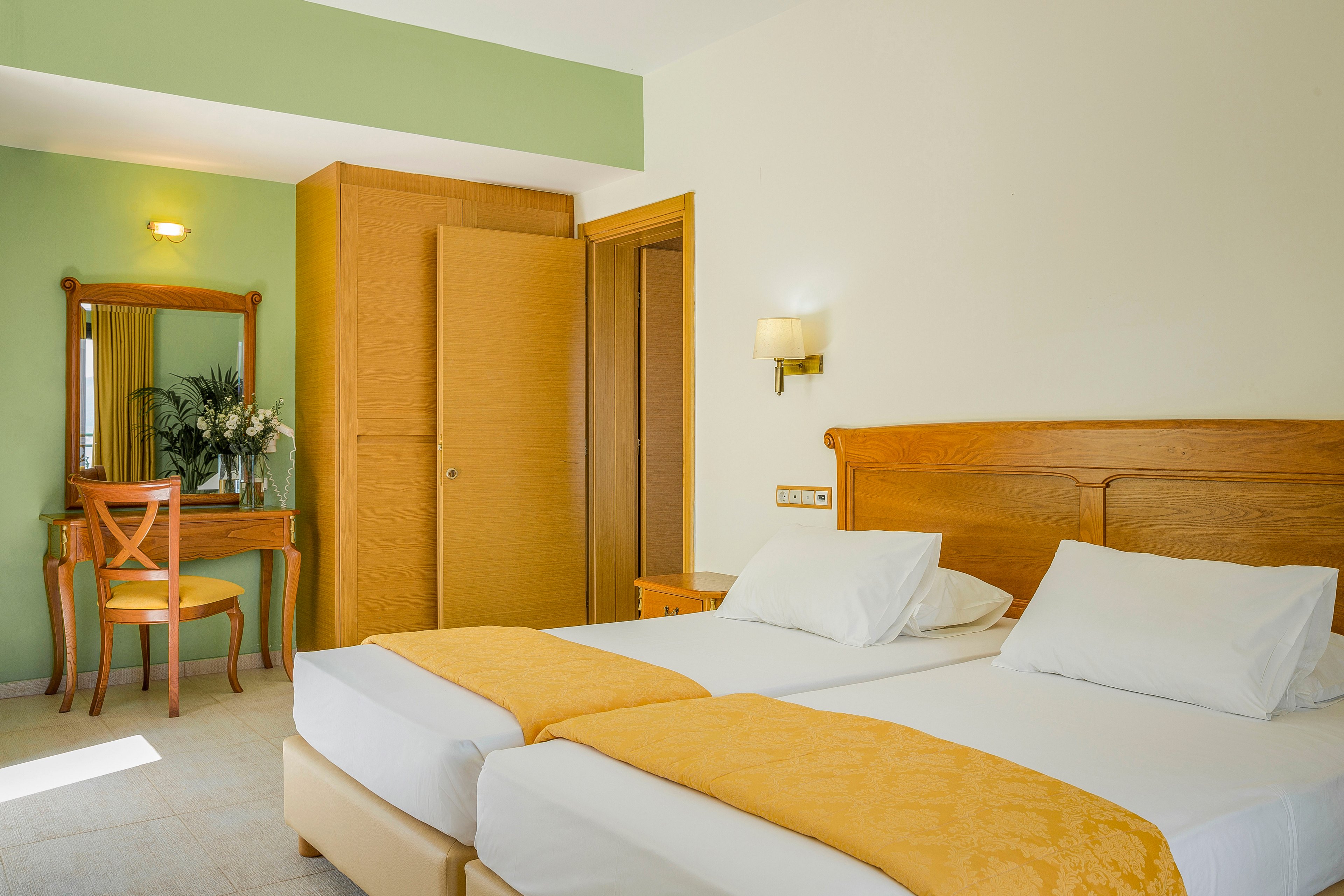 A sunlit hotel room features two neatly made twin beds with white linens and yellow throws. The space includes a wooden headboard, bedside wall lamp, and light-colored tiled floor. In the background, a wooden wardrobe and a dressing table with a mirror and chair add functionality. A vase with flowers on the vanity and soft green accent wall contribute to a fresh, welcoming atmosphere.