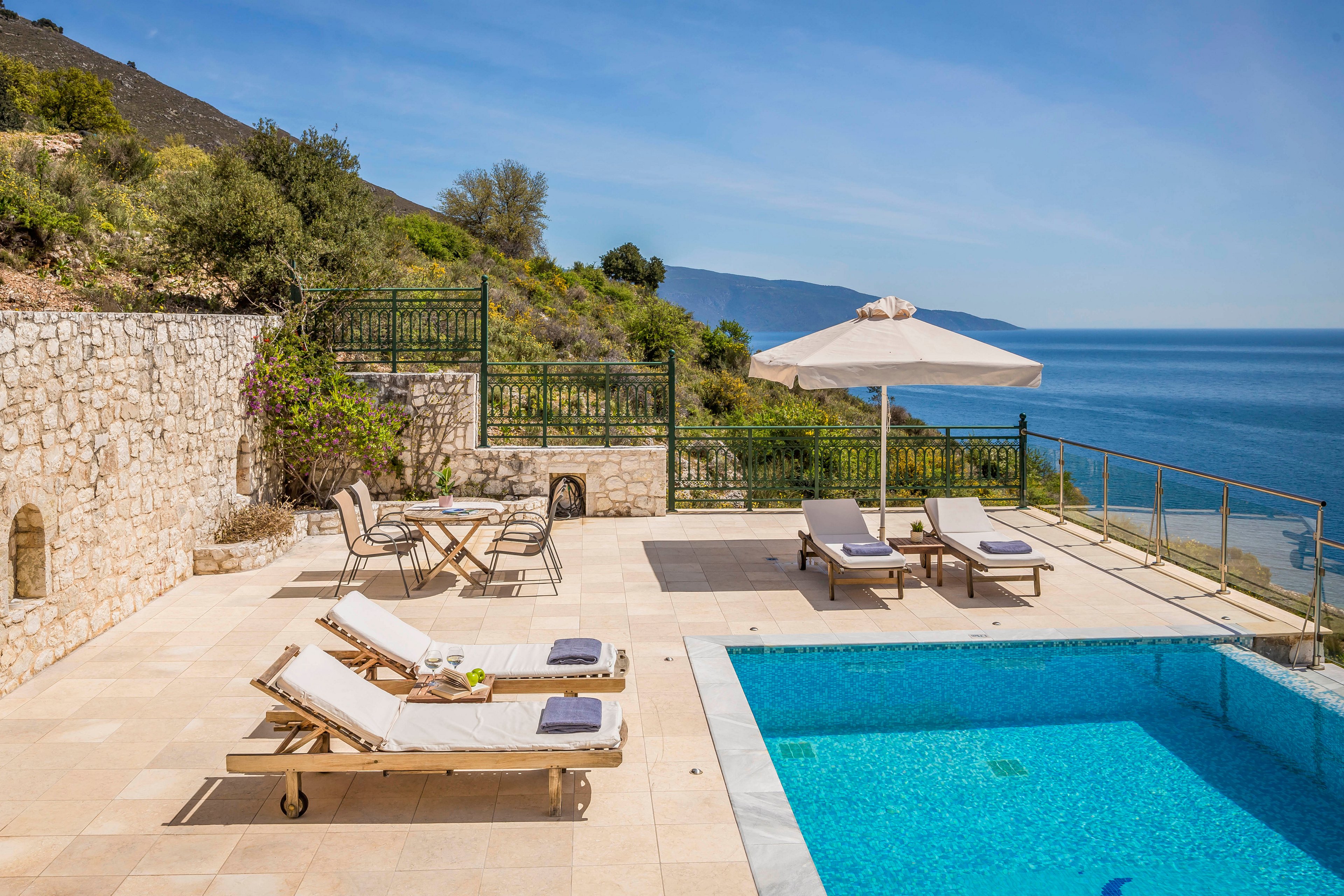 Sunny outdoor terrace with a bright blue swimming pool overlooks a calm sea and distant mountains. Wooden sun loungers with cushions and towels are arranged around the pool, alongside a large white parasol. A small table with chairs sits near a stone wall and wrought iron fence, surrounded by greenery. The clear sky and sparkling water create a relaxing vacation atmosphere.