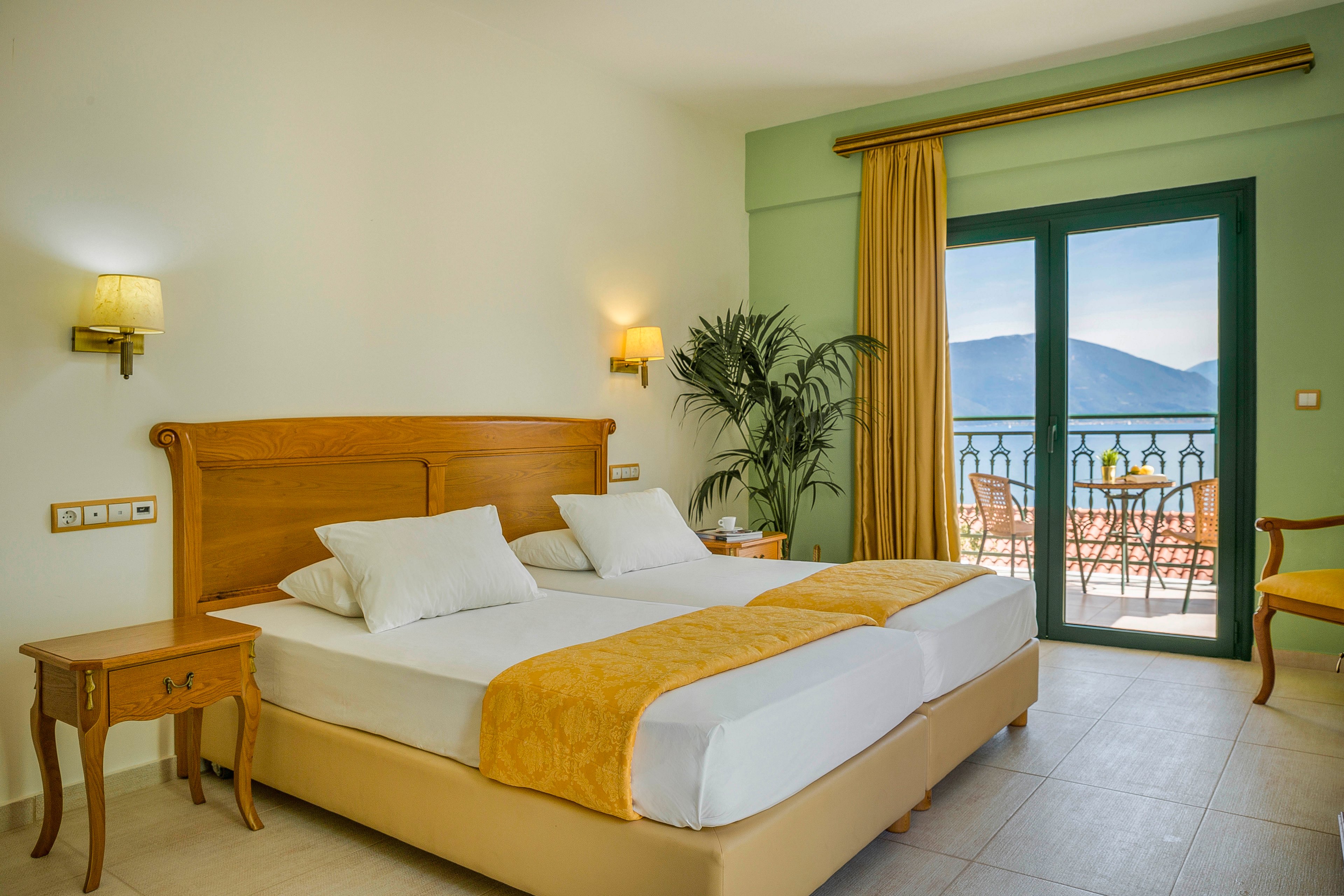 A tidy hotel bedroom features two single beds pushed together with white linens and yellow throws. The room is decorated in soft green and beige tones, with wooden furniture and wall-mounted lamps providing warm light. Large glass doors open onto a balcony with a table and chairs overlooking a calm sea and distant mountains. A tall indoor plant adds a touch of greenery to the relaxing coastal setting.
