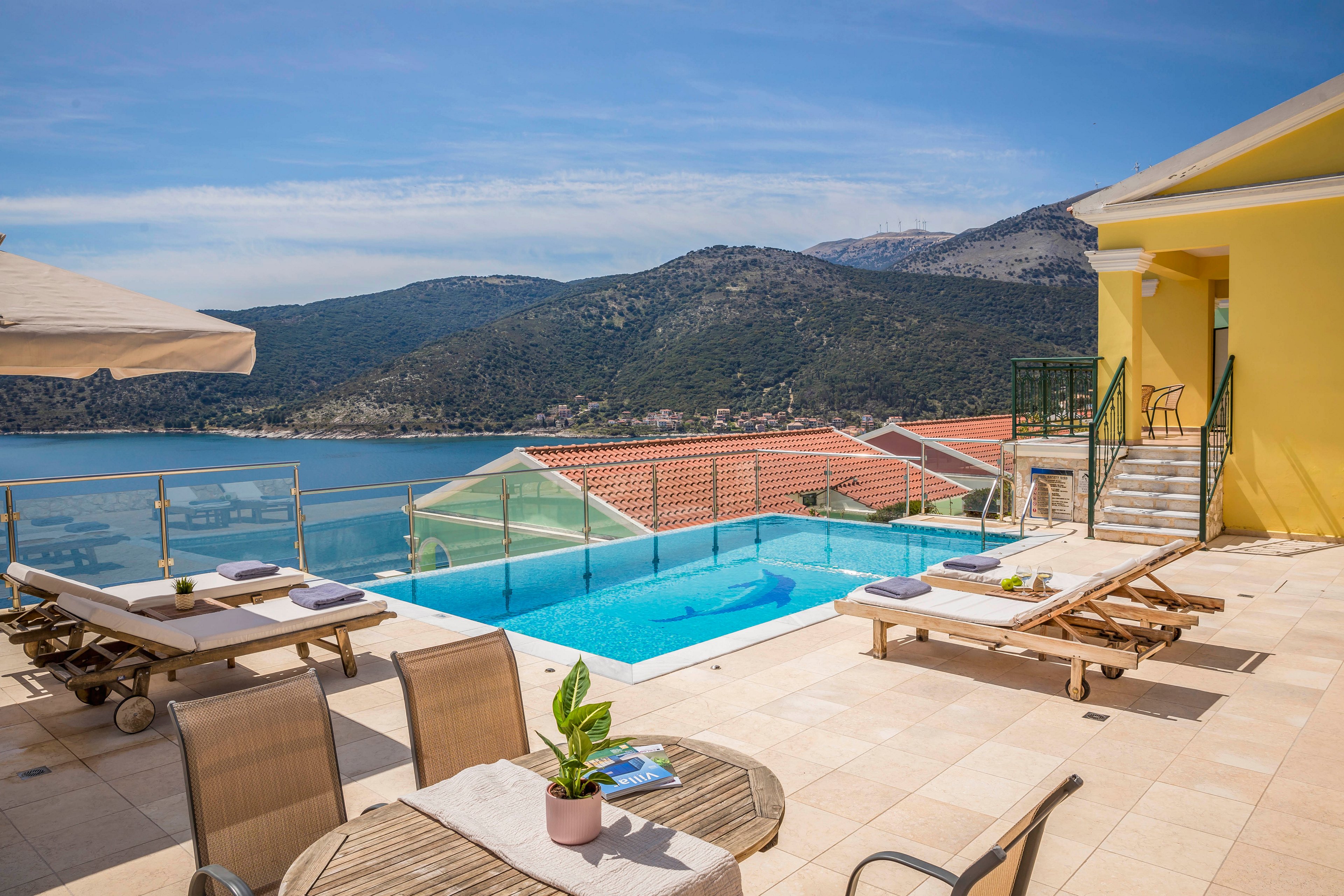 A bright villa terrace overlooks a clear blue infinity pool and a scenic bay. Lounge chairs and umbrellas are arranged around the pool, suggesting a relaxing vacation setting. In the foreground, a small table with chairs and a potted plant adds a cozy touch to the outdoor space. Hills and rooftops in the background complete the tranquil Mediterranean-style landscape under a clear sky.