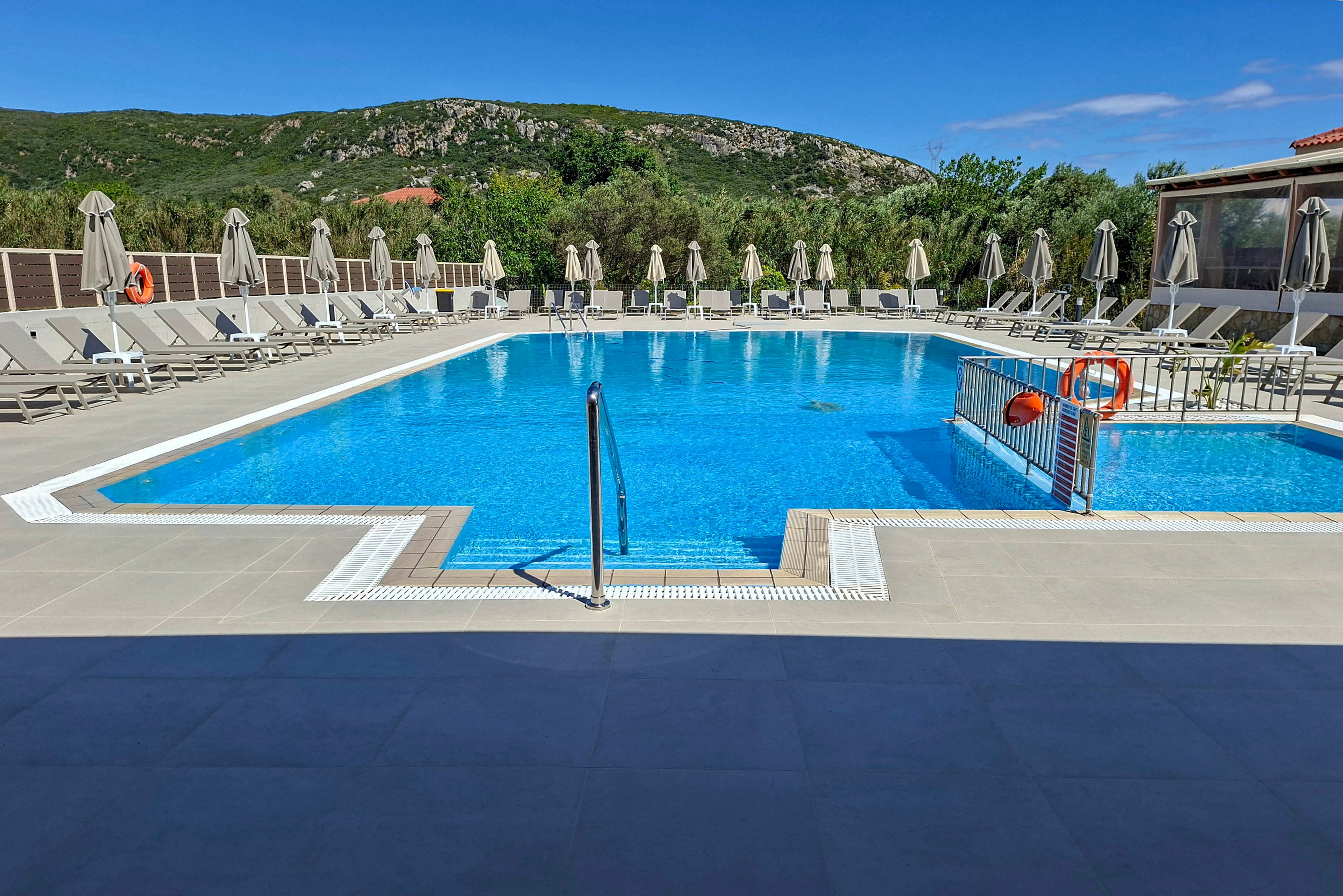 A clear blue outdoor swimming pool sits at the center of a quiet resort. Numerous sun loungers and closed umbrellas line the poolside terrace under a bright blue sky. A metal handrail leads into the water, and safety equipment such as life rings is visible near the pool edge. Green hills and trees form a scenic natural backdrop beyond the resort buildings.