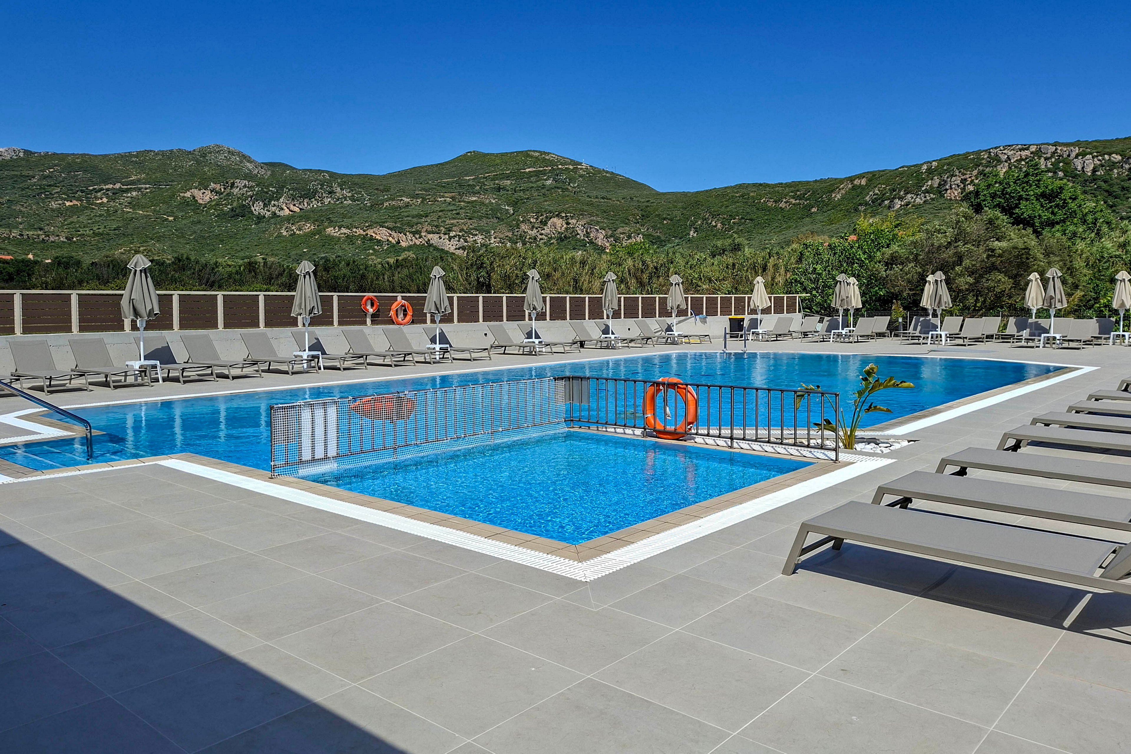An outdoor swimming pool sits empty at a modern resort with sun loungers neatly arranged around the tiled deck. Beige umbrellas and safety rings line the poolside, while metal railings separate the shallow and deep sections. Green hills and mountains form a scenic backdrop under a clear blue sky. The scene feels calm and inviting, suggesting a peaceful vacation setting.