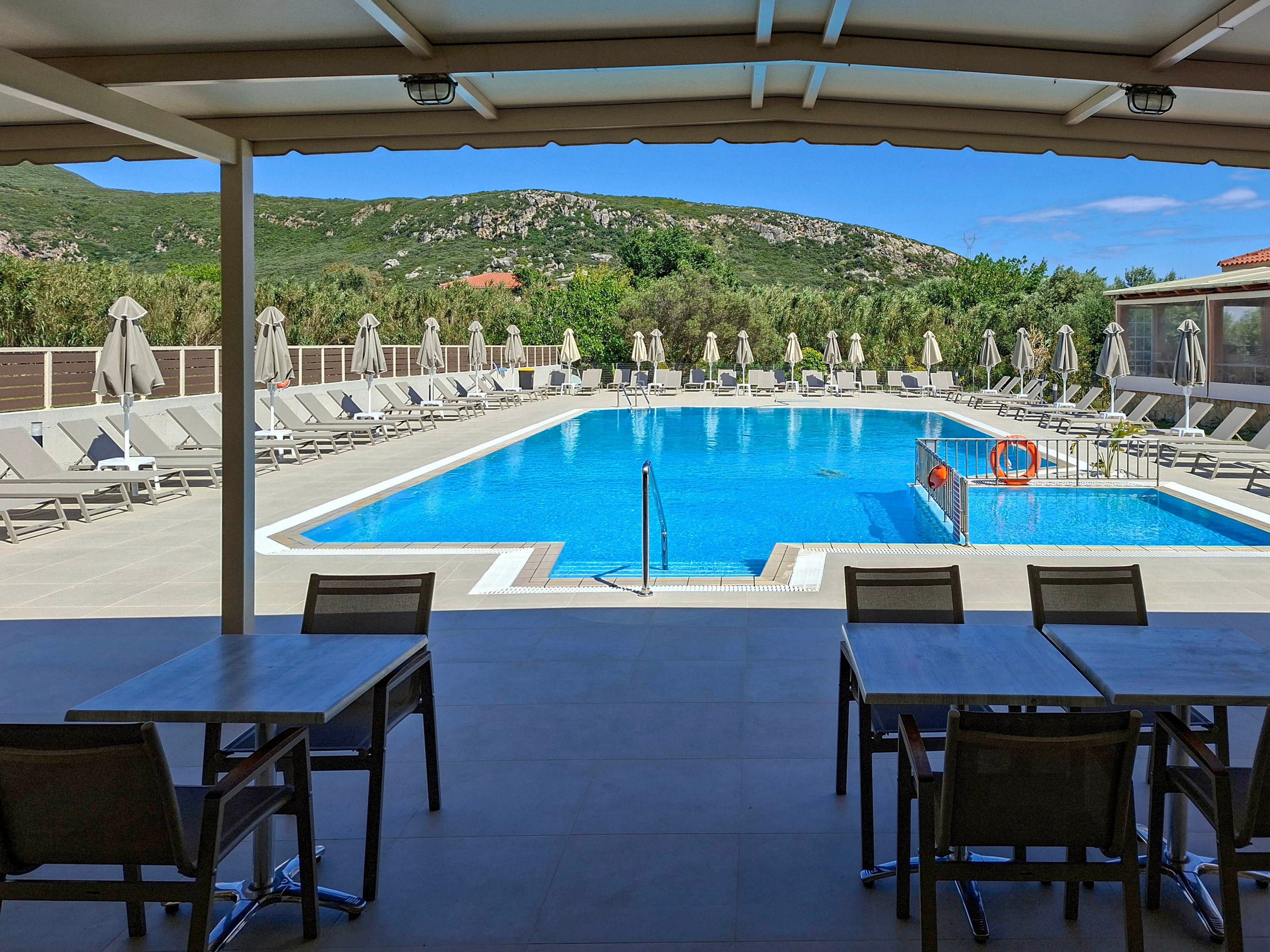 A large outdoor swimming pool is surrounded by rows of sun loungers and umbrellas at a resort. The scene is viewed from a shaded terrace with empty tables and chairs in the foreground. Clear blue water reflects the bright sky, while a safety ring and metal ladder are visible at the poolside. Green hills and rocky mountains form a scenic backdrop under sunny weather.