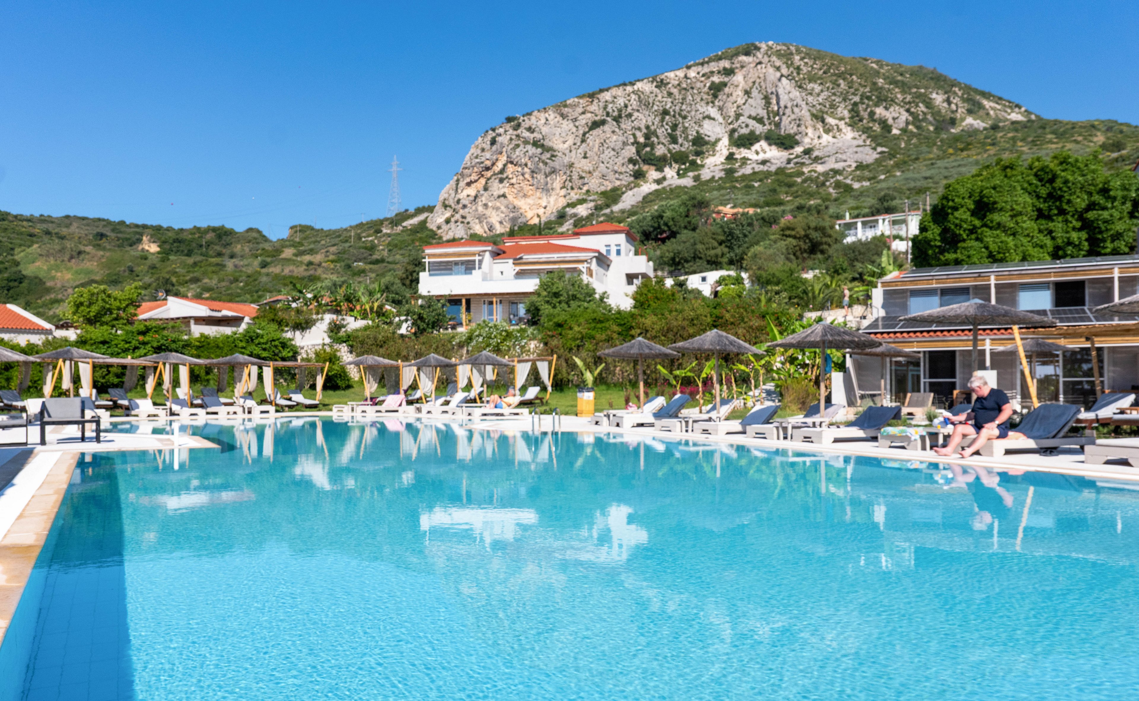 A large outdoor swimming pool reflects the clear blue sky at a hillside resort. Sun loungers and parasols line the poolside, where a few guests relax in the sun. White villas with red roofs are nestled among lush greenery in the background. A rocky mountain rises behind the buildings, creating a scenic vacation setting.