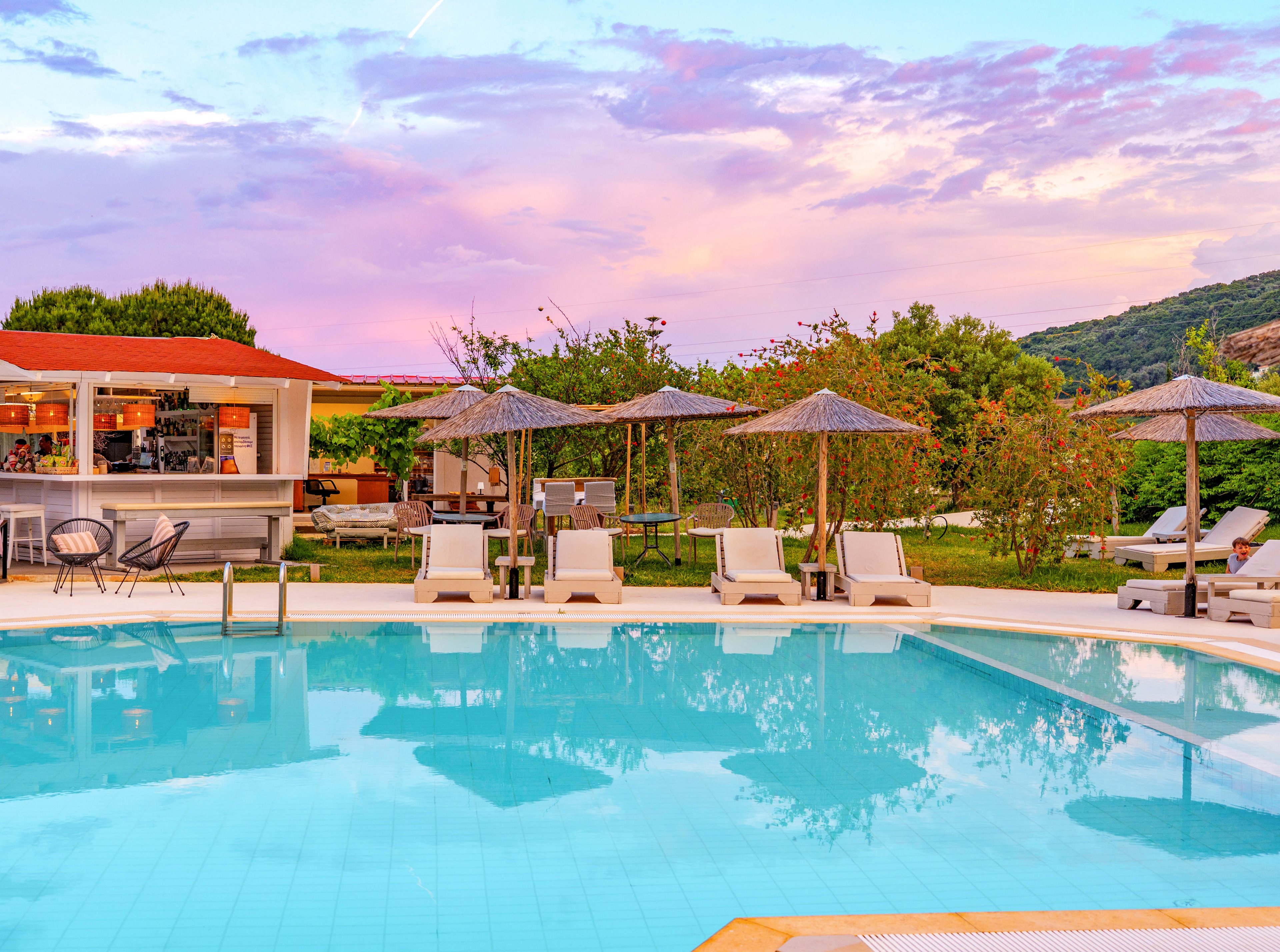 An inviting resort swimming pool reflects the pastel colors of a vibrant sunset sky. Sun loungers and straw parasols line the poolside, creating a relaxing vacation atmosphere. A modern outdoor bar with seating is visible in the background, surrounded by lush greenery and low hills. The calm water and warm evening light emphasize a tranquil, luxurious holiday setting.