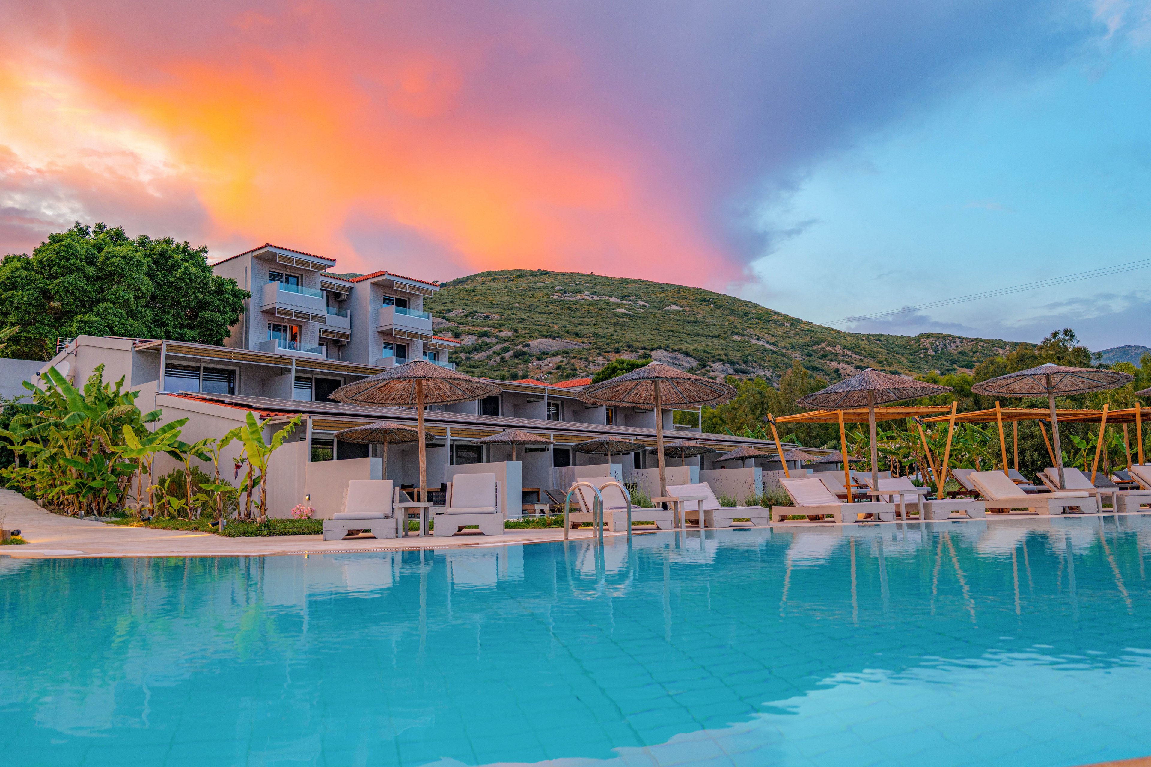 A contemporary resort features a large outdoor swimming pool reflecting the vivid colors of a sunset sky. White lounge chairs and umbrellas line the poolside, creating a relaxing vacation atmosphere. Low-rise hotel buildings sit against a backdrop of green hills and trees. The calm water and warm evening light emphasize a tranquil, luxurious holiday setting.