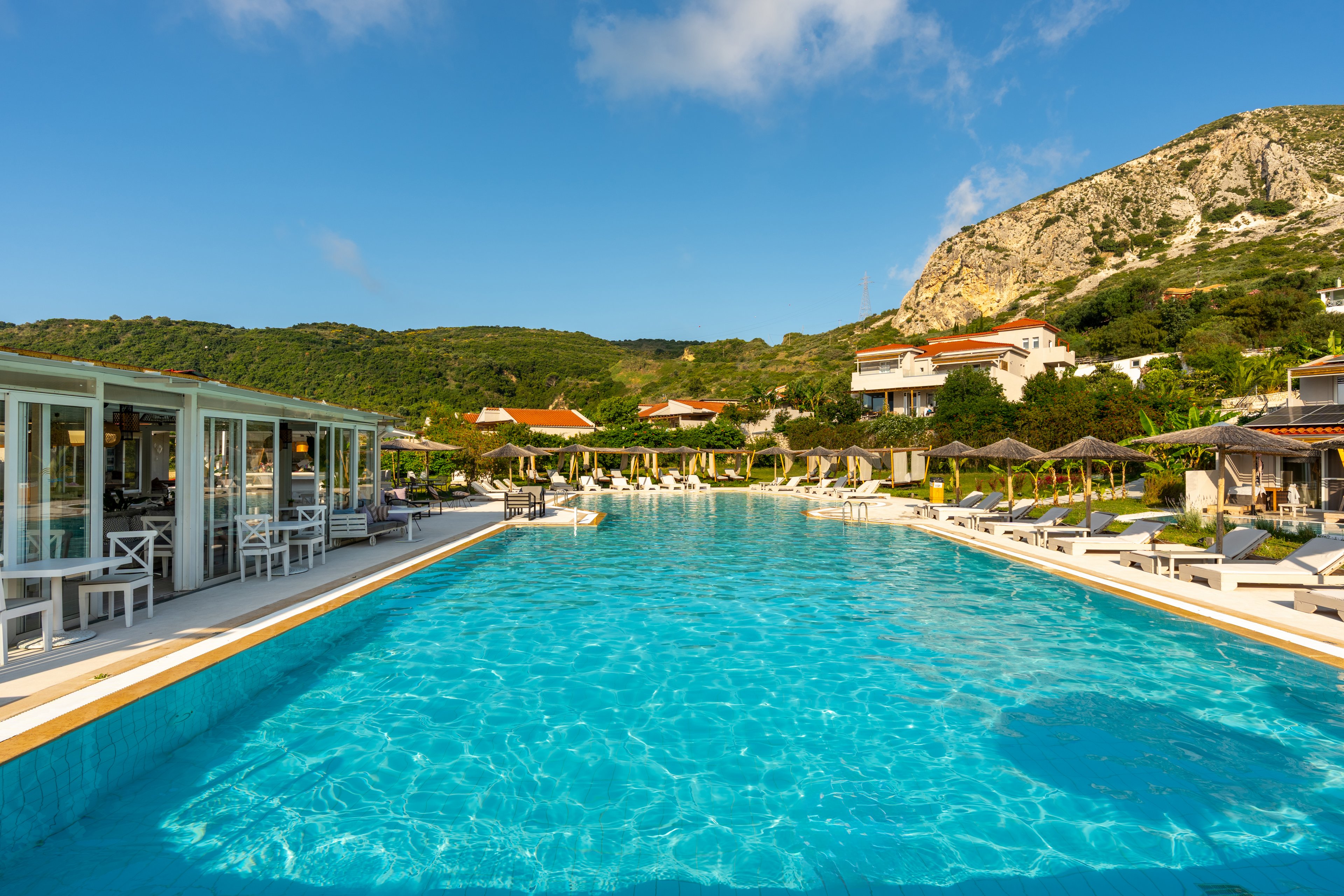 A large turquoise swimming pool stretches across the foreground of a sunny resort. Sun loungers and umbrellas line the poolside, with open-air seating and glass-fronted buildings on the left. Green hills and rocky mountains rise behind the property, dotted with red-roofed houses. The clear blue sky and bright reflections in the water create a relaxed vacation atmosphere.