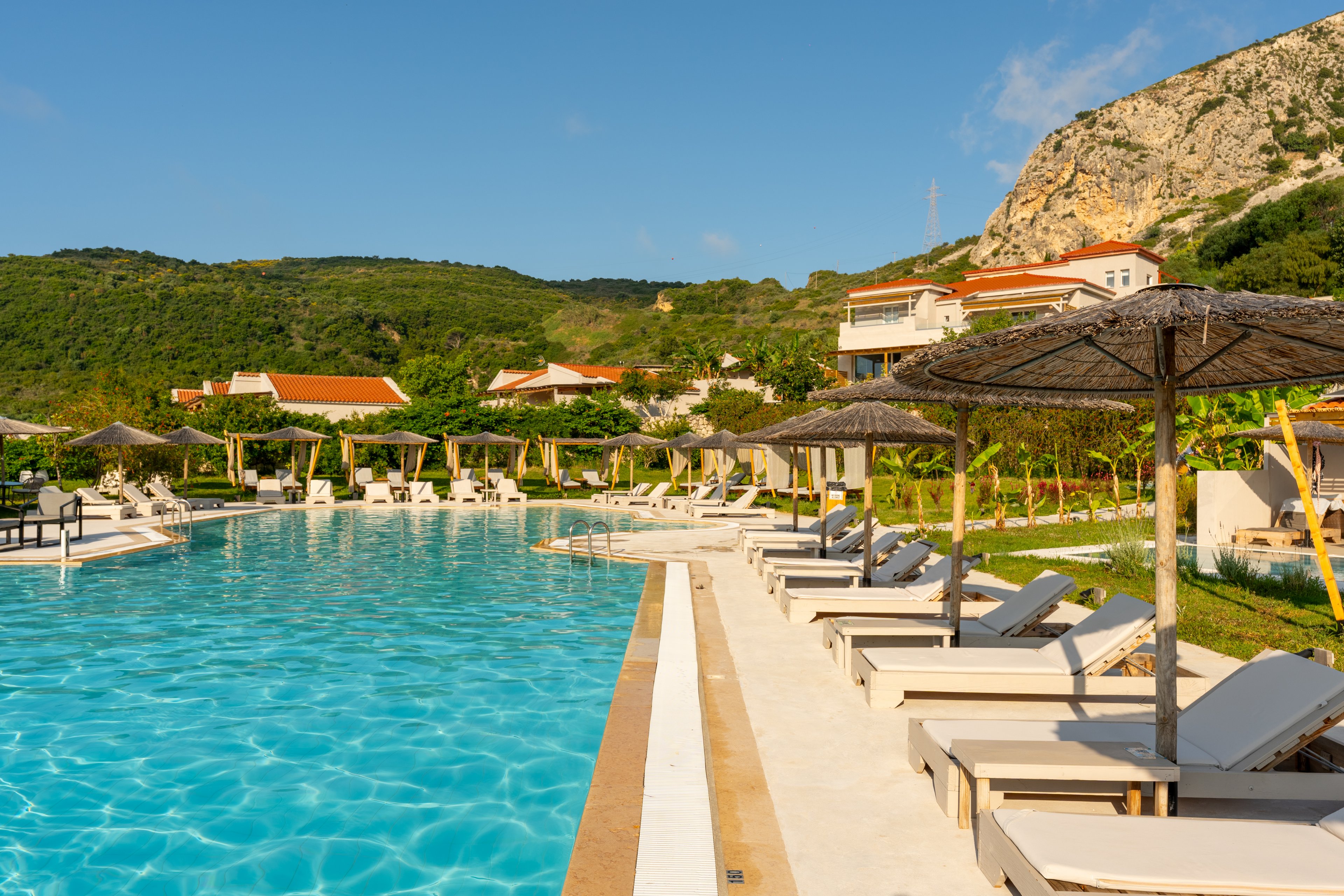 A large outdoor swimming pool is surrounded by rows of white sun loungers and straw parasols at a hillside resort. The scene is set on a bright sunny day with clear blue water reflecting the sky. Green hills and rocky cliffs rise in the background, dotted with red-roofed buildings. The overall atmosphere is calm and inviting, suggesting a relaxing vacation destination.
