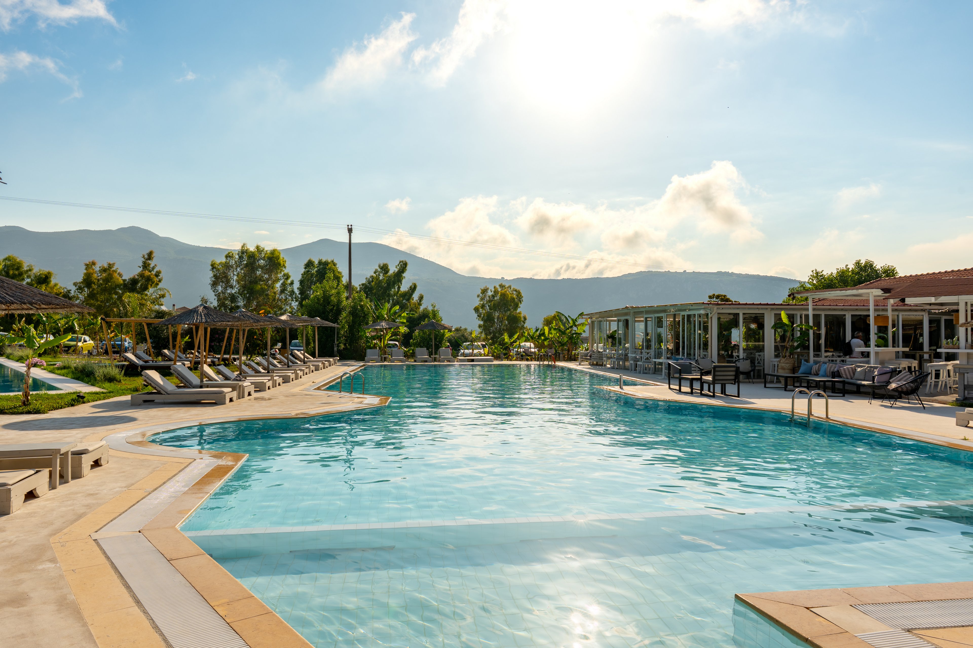 Large outdoor swimming pool at a resort is shown under bright afternoon sunlight. Sun loungers and umbrellas line the poolside, with modern open-air buildings and terraces in the background. Green trees and plants surround the area, leading to distant mountains under a partly cloudy sky. The calm water and warm light create a relaxed vacation atmosphere.