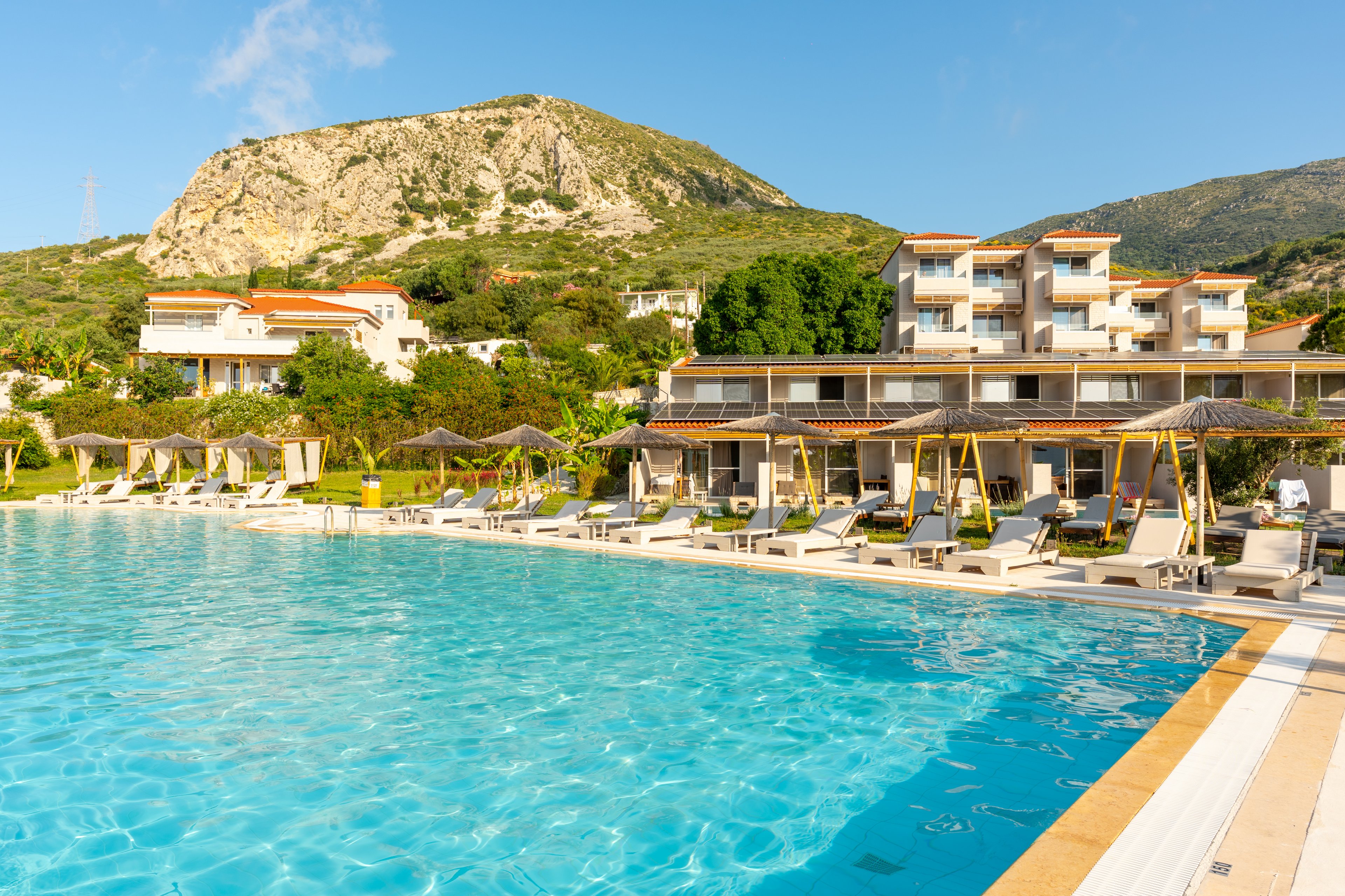 A large turquoise swimming pool stretches across the foreground of a modern resort complex. Sun loungers and umbrellas line the poolside, suggesting a relaxing vacation atmosphere. Multi-story buildings with balconies sit amid lush greenery at the base of a rocky mountain. The scene is brightly lit by warm sunlight under a clear blue sky, evoking a tranquil Mediterranean or coastal getaway.