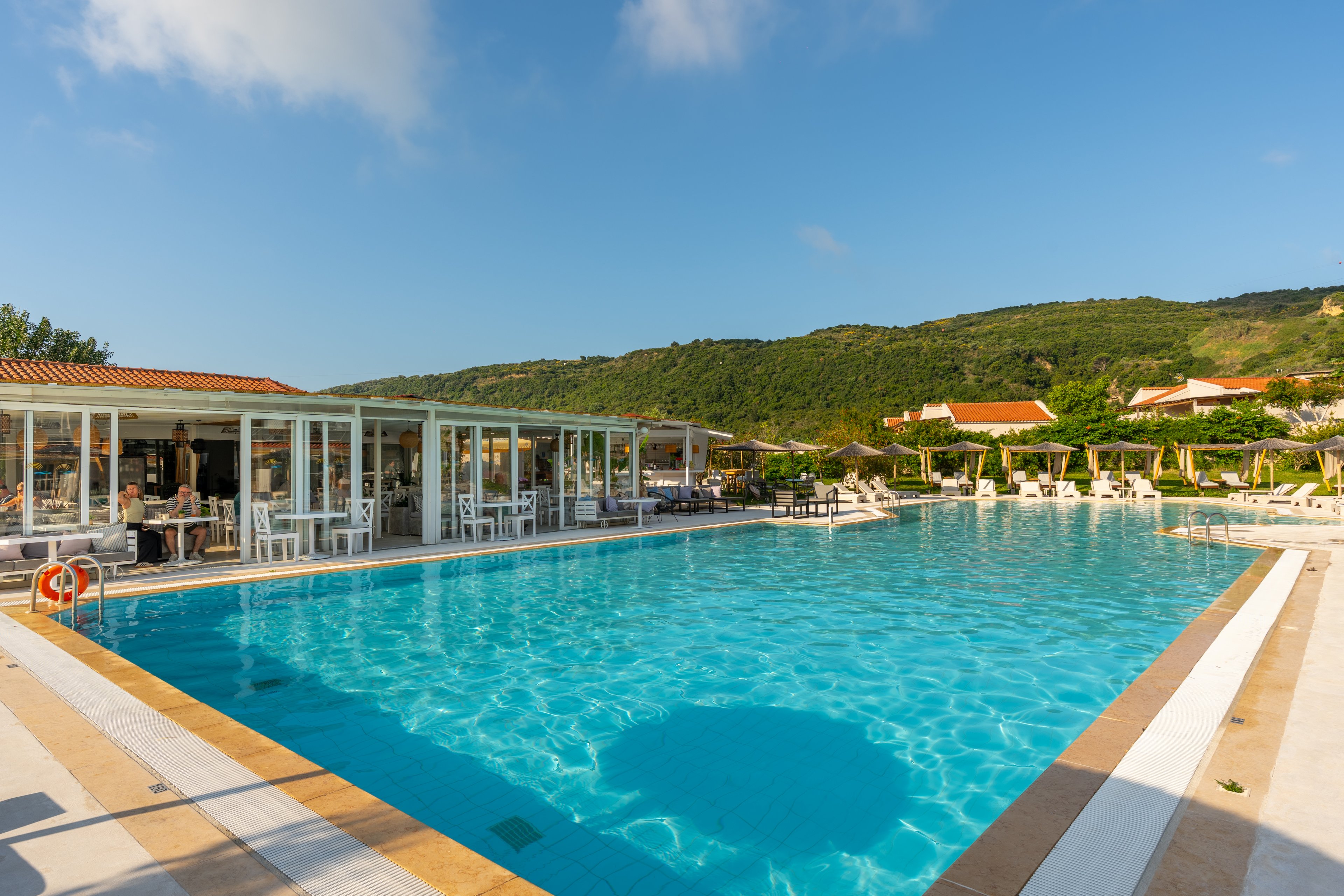 A large outdoor swimming pool reflects the bright blue sky at a resort nestled below green hills. Sun loungers and umbrellas line the poolside, with a glass-fronted building housing a bar or restaurant on one side. A few people relax near the water, enjoying the warm, sunny weather. The scene conveys a tranquil vacation atmosphere with clear water and inviting leisure facilities.