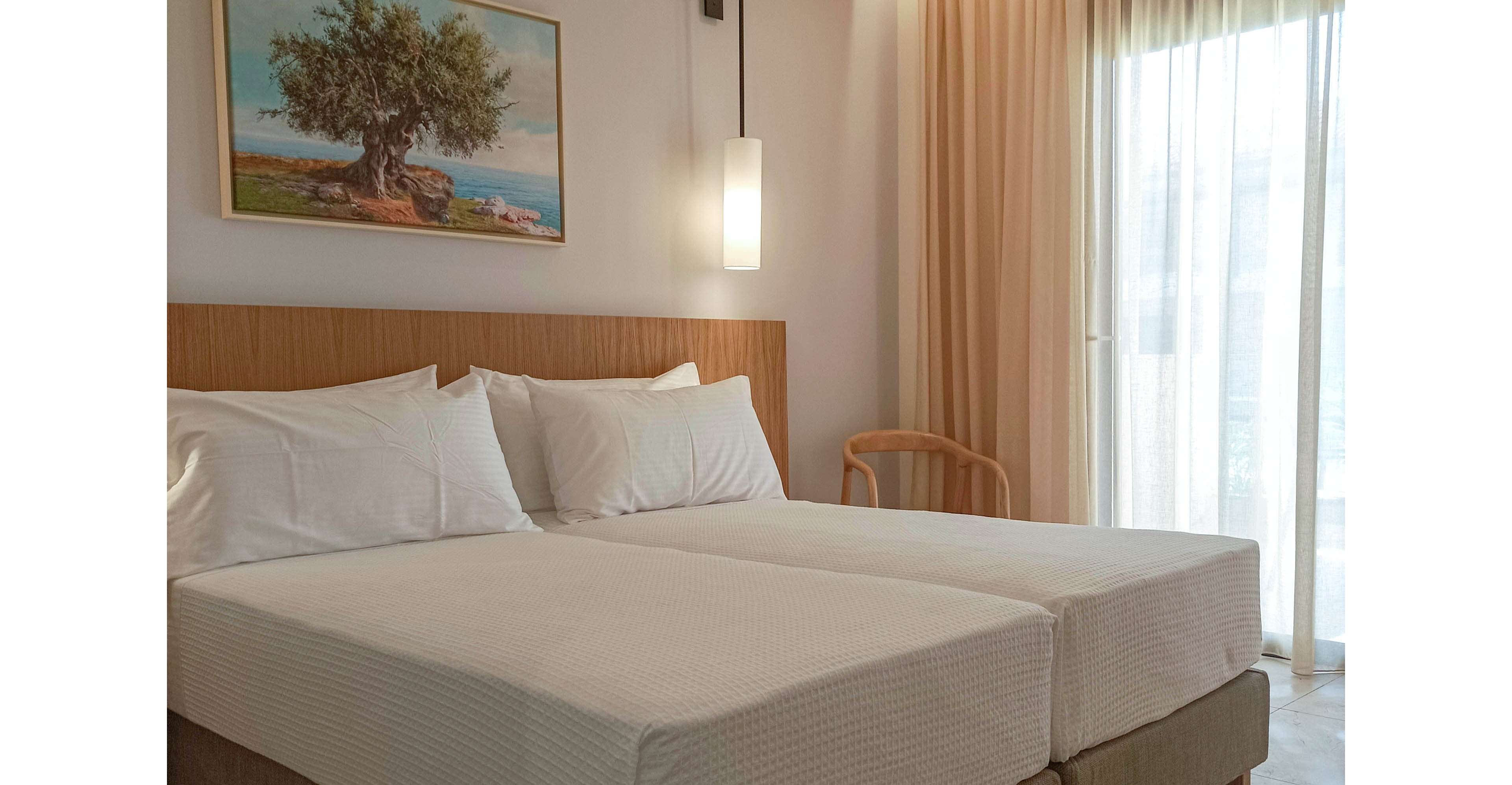 A contemporary hotel bedroom features two neatly made twin beds with white linens and a wooden headboard. Soft natural light enters through a large window covered with sheer beige curtains. A modern pendant light hangs above the bedside area, and a simple chair sits in the corner. A framed landscape painting of a tree and sea view decorates the wall, enhancing the calm and inviting atmosphere.