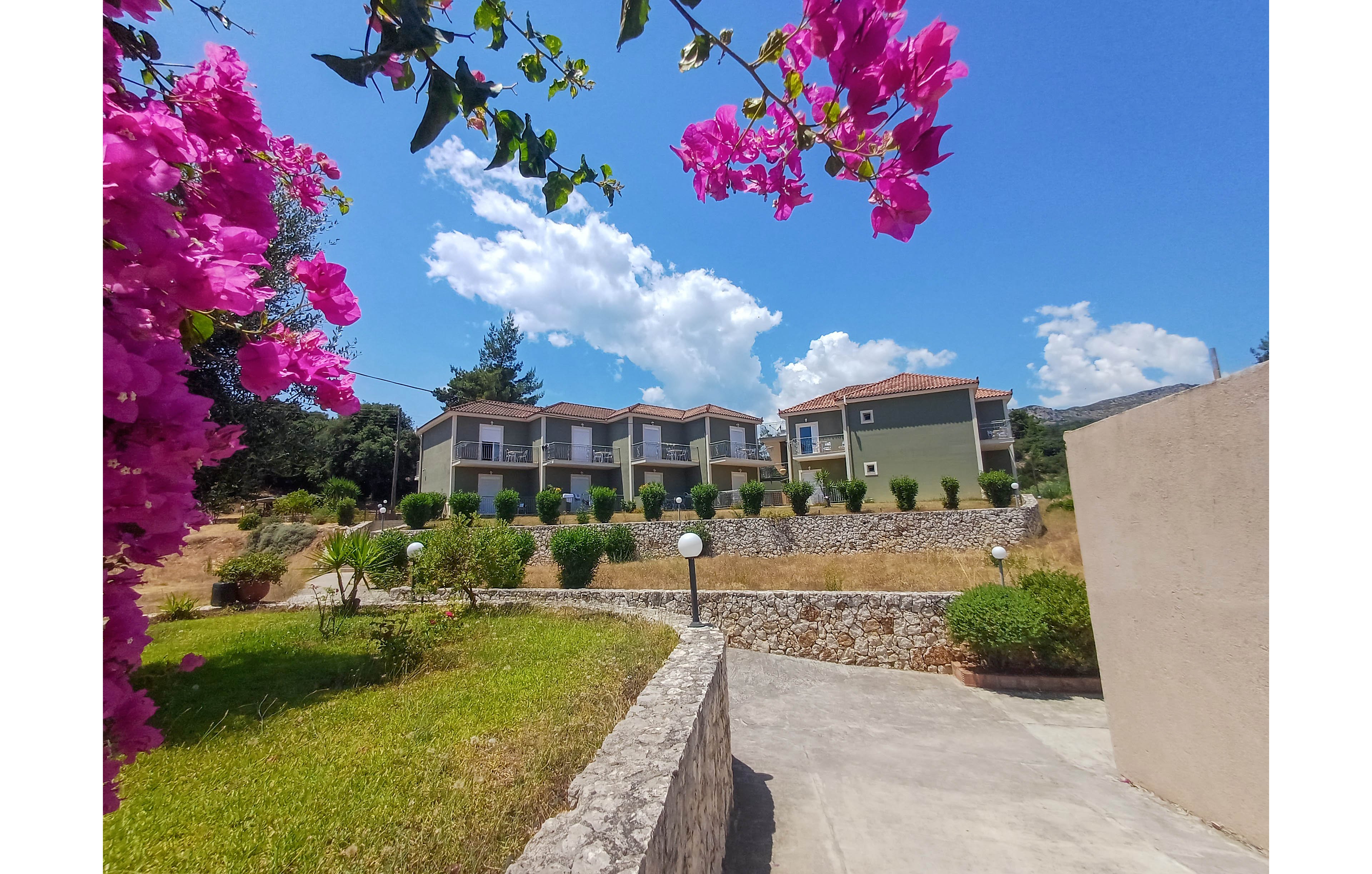 A row of modern, light-colored apartment buildings stands on a terraced hillside under a bright blue sky with scattered clouds. A concrete path leads toward the residences, bordered by stone walls and neatly trimmed green shrubs. Vibrant pink flowers frame the foreground, adding a colorful accent to the sunny outdoor scene. The overall atmosphere is calm and inviting, suggesting a vacation or residential complex in a warm climate.