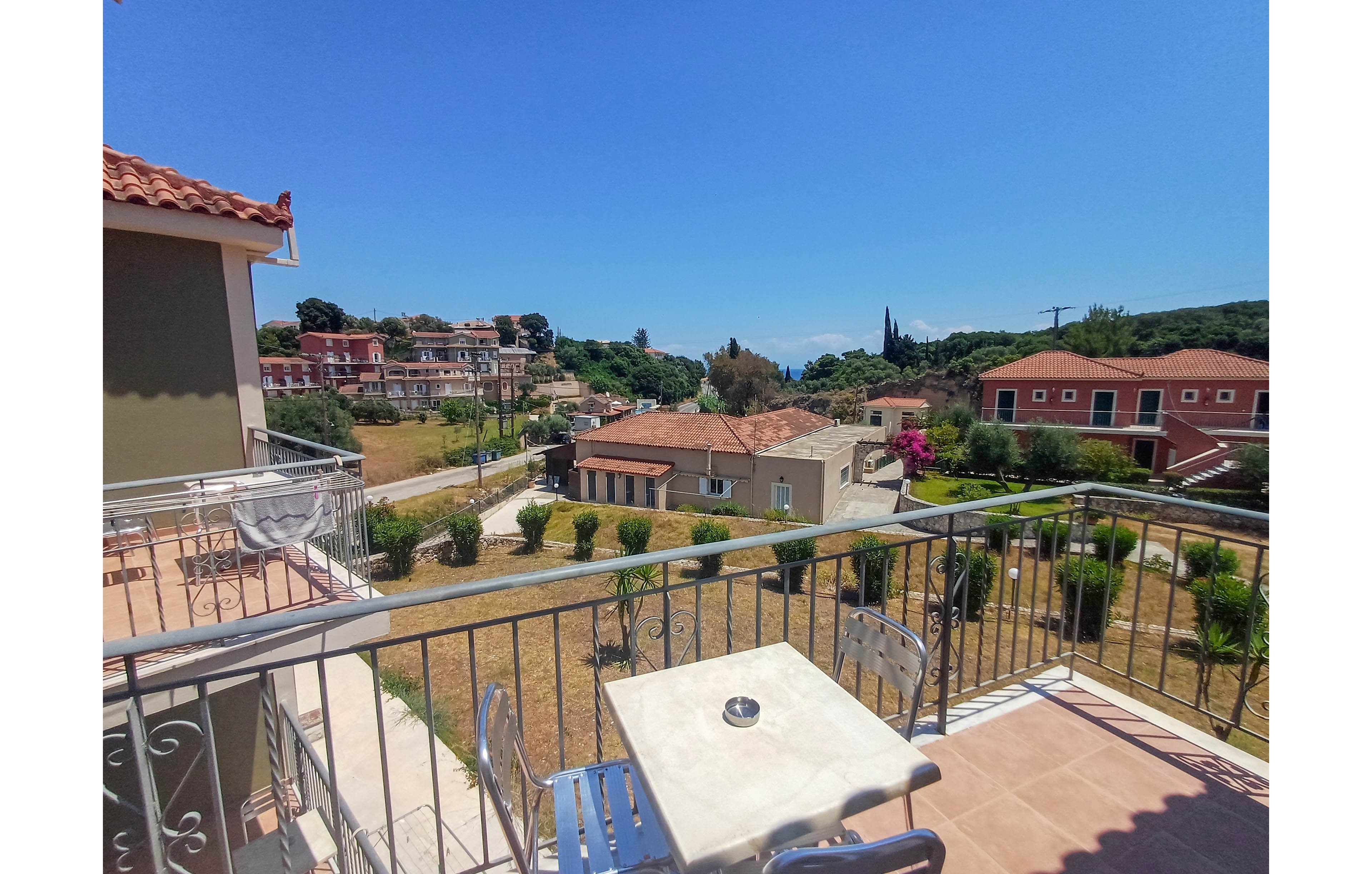 A small balcony with metal chairs and a white table overlooks a quiet residential area. Terracotta-roofed houses and low buildings are surrounded by dry grass, shrubs, and palm trees. In the distance, lush green hills and a glimpse of blue sea meet a clear, bright sky. The scene suggests a warm, peaceful day in a coastal Mediterranean town.