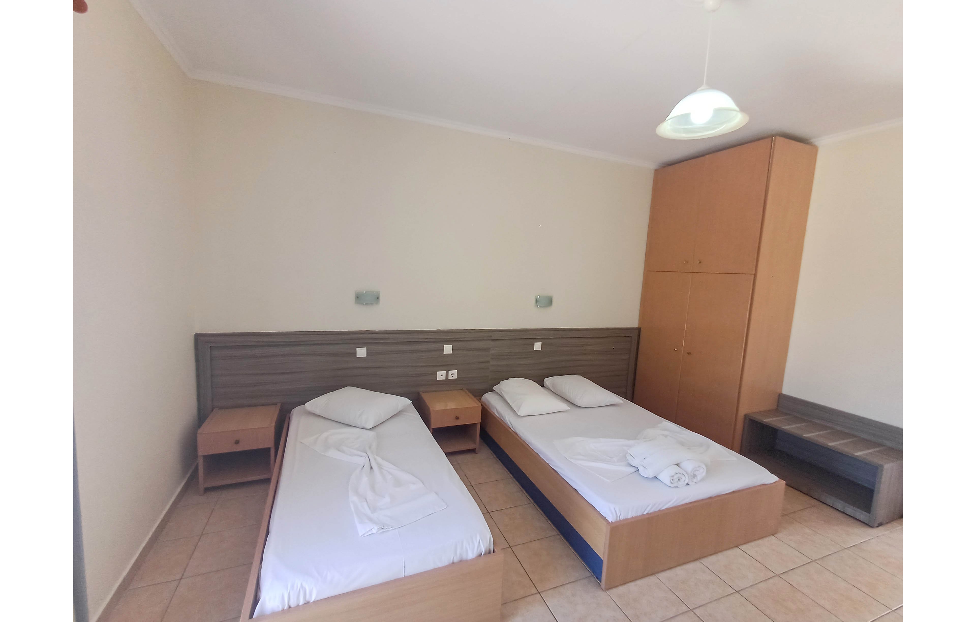 The image shows a modest hotel or guest room with two single beds placed side by side. Each bed has white sheets and pillows, with towels neatly folded on one of the beds. Wooden bedside tables, a wardrobe, and a bench complement the neutral tiled floor and light-colored walls. The room is brightly lit, creating a clean and functional atmosphere suitable for budget travel or short stays.