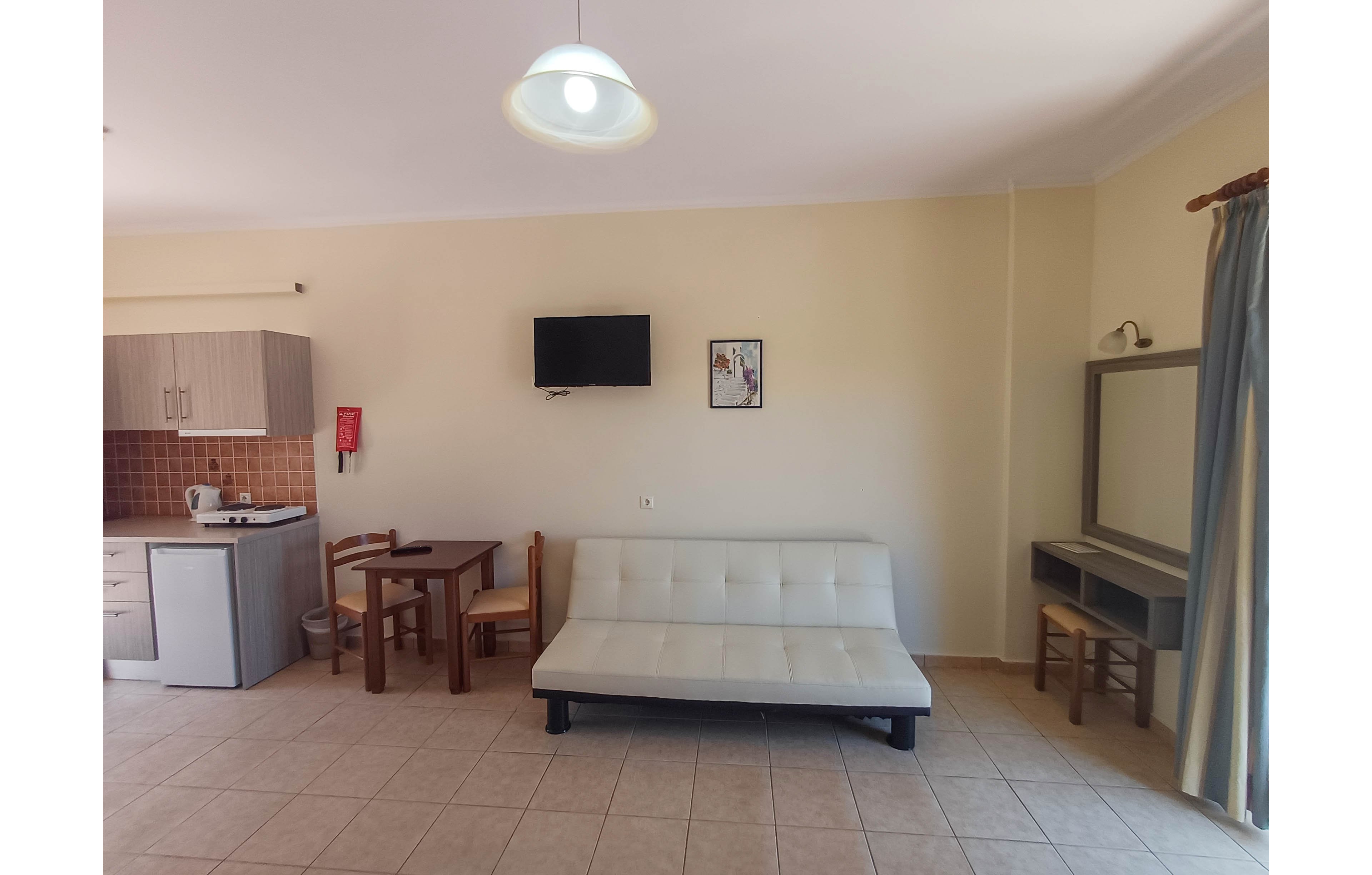 A compact studio living area features a white futon sofa against a light-colored wall. A small wall-mounted television and framed artwork hang above the seating. On the left, a kitchenette with cabinets, sink, and tiled backsplash is accompanied by a wooden dining table and chairs. On the right, a dressing table with mirror sits near a window with curtains, allowing natural light into the tiled room.