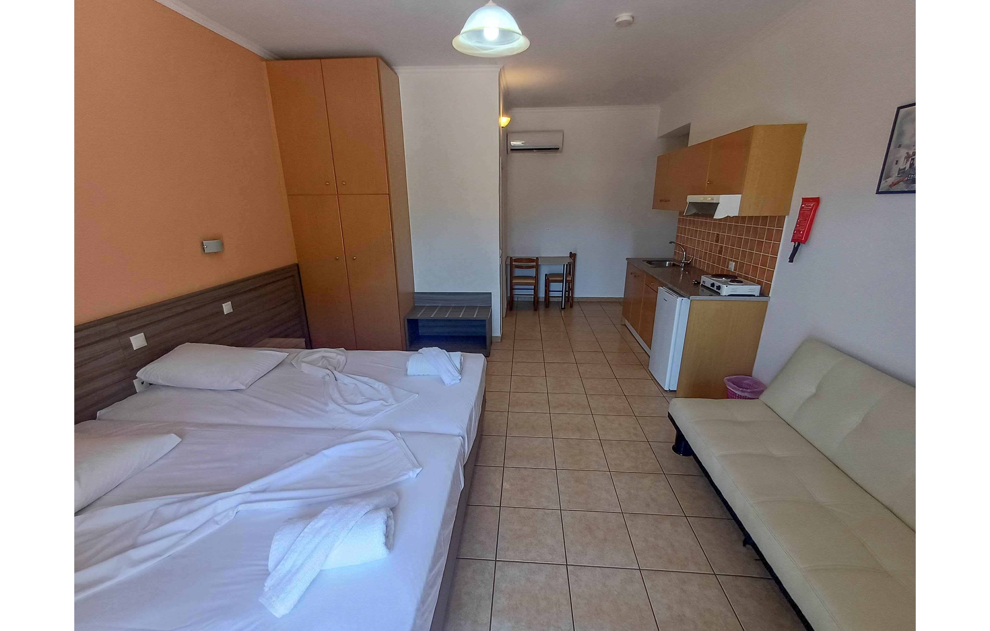 A small studio room features two beds pushed together with white linens and towels. The space includes a kitchenette with wooden cabinets, tiled backsplash, sink, stovetop, and a compact refrigerator. A beige sofa sits opposite the beds, and a small table with two chairs is placed near the back wall. The room has tiled flooring, an air conditioning unit, and simple overhead lighting, creating a clean and functional vacation rental atmosphere.