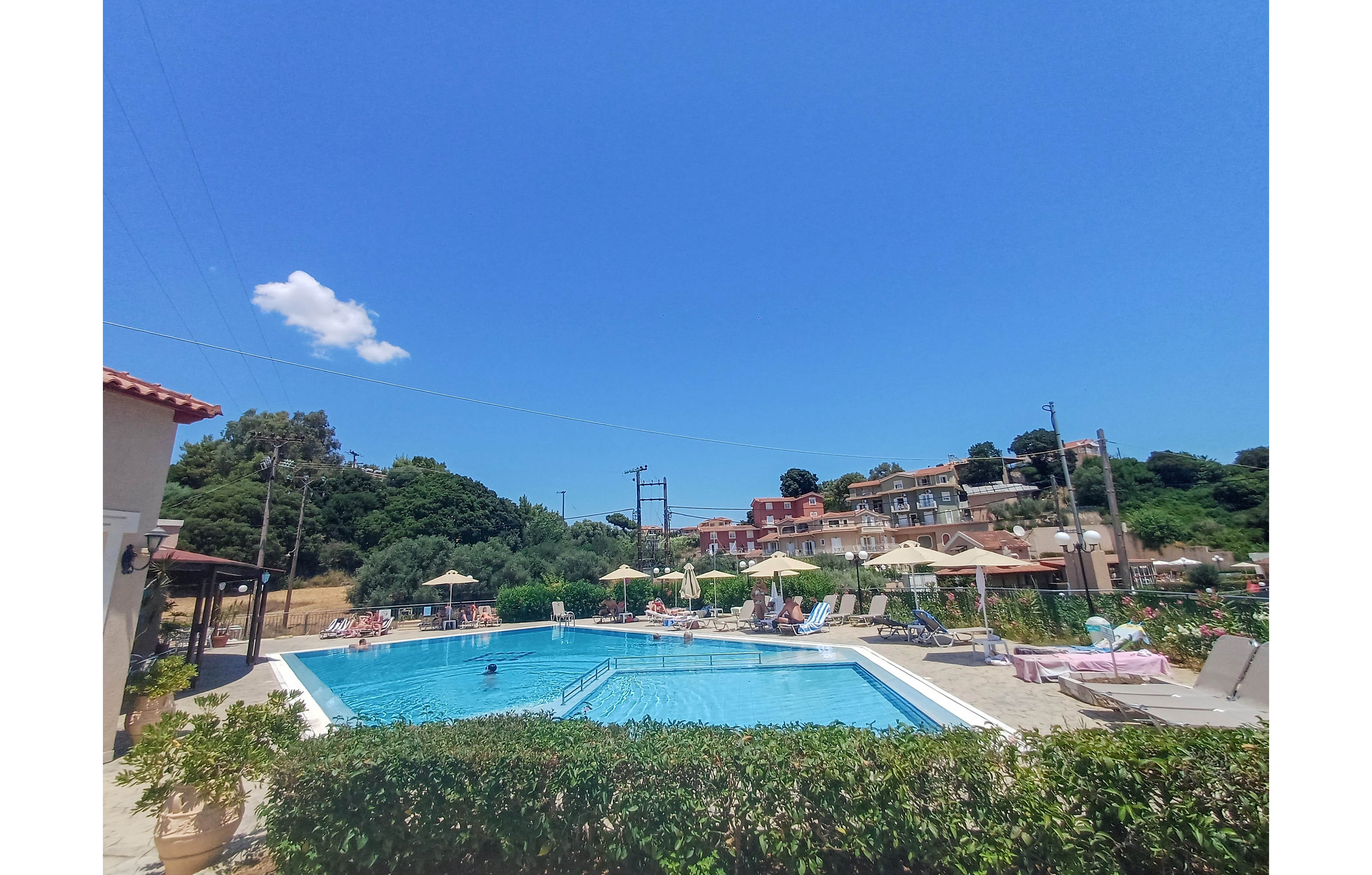 An outdoor swimming pool is surrounded by sun loungers and umbrellas at a hillside resort. A few people relax by the water while one person swims in the clear blue pool. Green shrubs frame the foreground, and multi-story buildings rise behind the pool area. The sky above is bright and cloudless except for a single small white cloud, creating a warm vacation atmosphere.