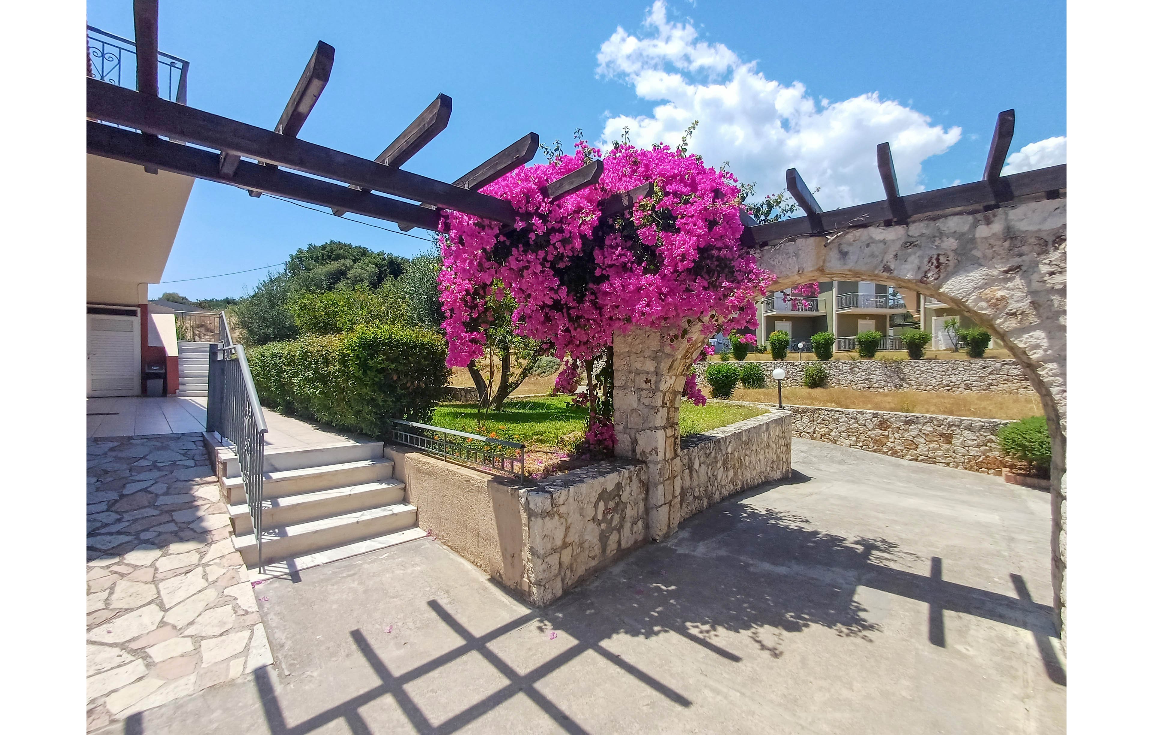 A sunlit courtyard features a stone arch covered in bright pink bougainvillea flowers. Steps lead up from a paved walkway to a raised garden area with neatly trimmed bushes and trees. In the background, residential buildings and terraced stone walls are visible under a blue sky with scattered white clouds. Strong sunlight casts shadows from the wooden pergola beams across the ground, emphasizing the warm, Mediterranean atmosphere.