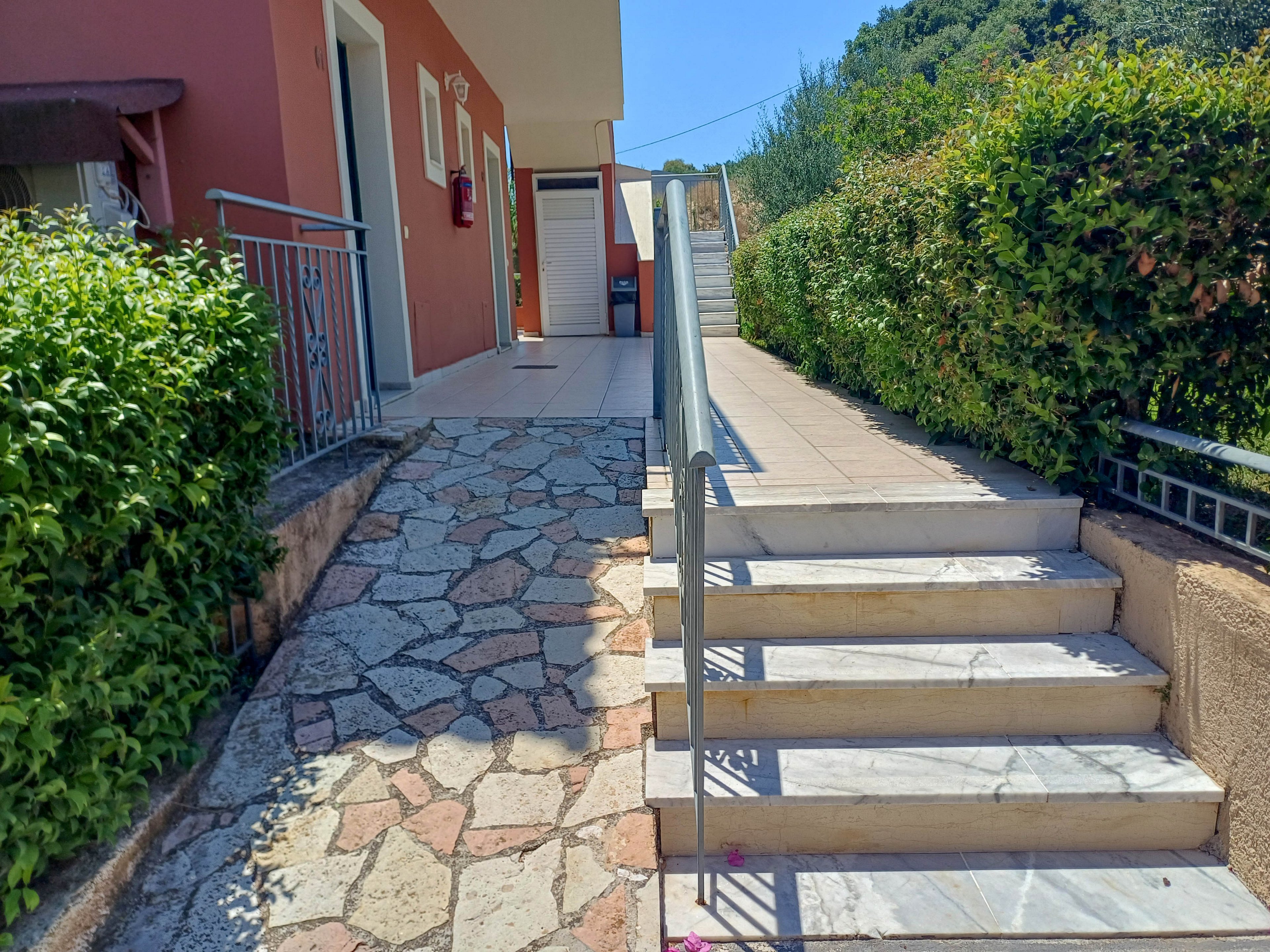 An exterior stairway with a metal handrail leads up beside a stone walkway and tiled ramp. The scene is part of a residential or vacation building with orange walls and doors visible along the corridor. Dense green hedges border the stairs and path, and sunlight creates strong shadows on the ground. Trees and vegetation are visible in the background under a clear blue sky.