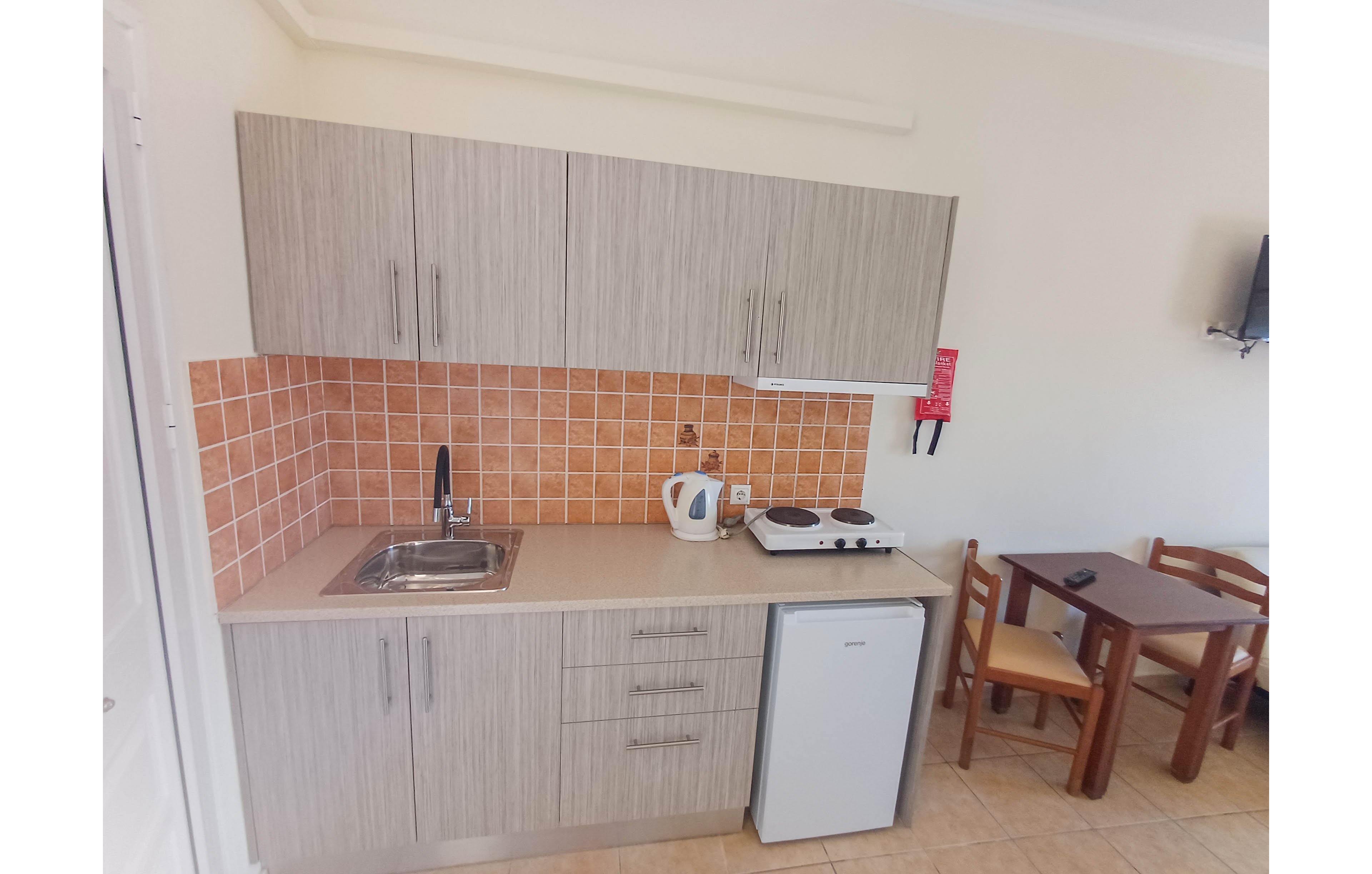 A small studio kitchenette features light wood cabinets and an integrated stainless steel sink. Orange tiled backsplash contrasts with the neutral countertop, where a white electric kettle and a two-burner hot plate are placed. A compact under-counter refrigerator sits beneath the counter, and a simple wooden dining table with two chairs stands nearby. The space has a clean, minimal look suitable for short-term stays or small apartments.