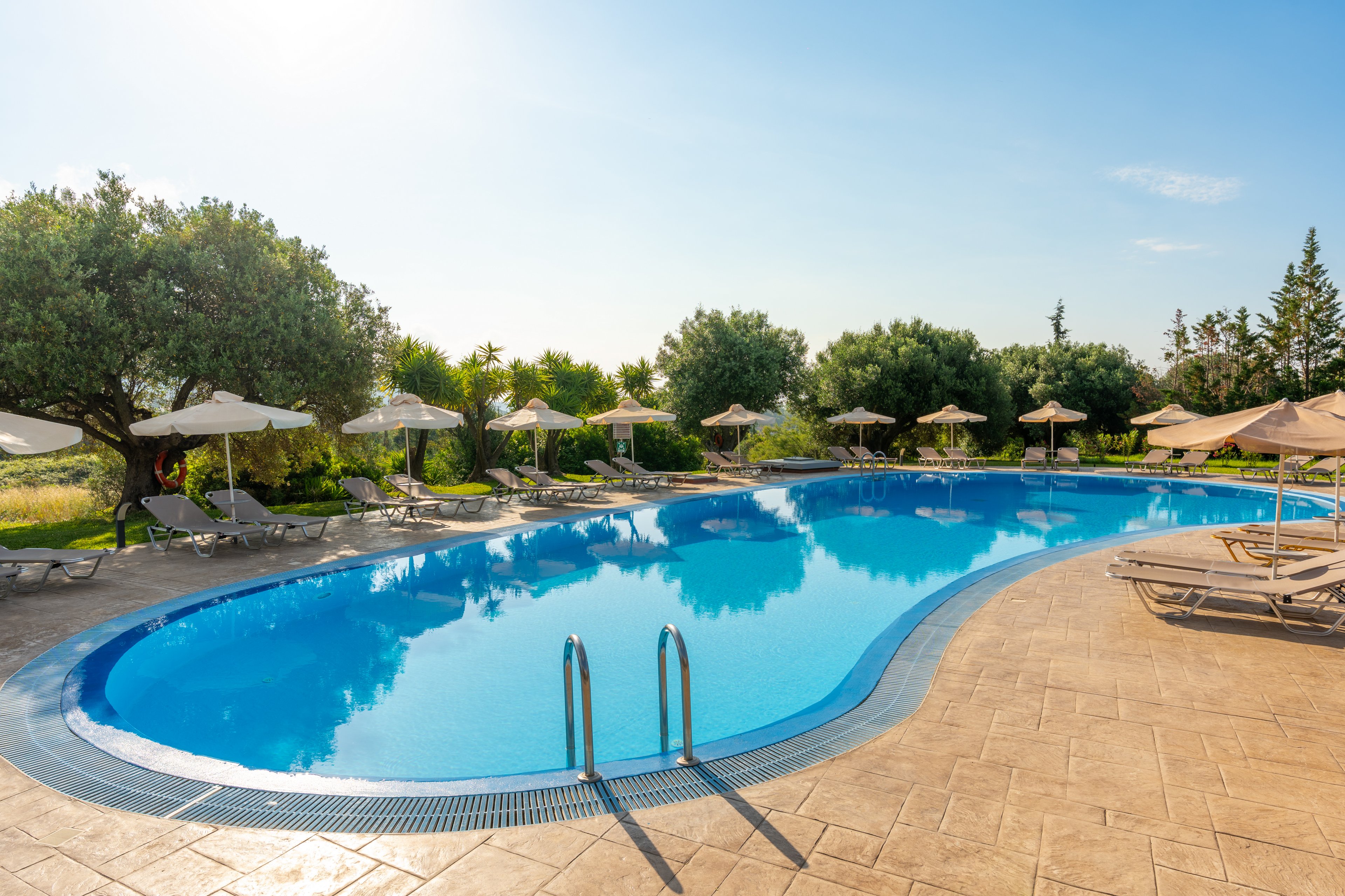 An outdoor swimming pool curves through a sunlit resort setting. Sun loungers and parasols line the tiled pool deck, inviting relaxation. Green trees and landscaped gardens surround the area under a clear blue sky. The calm water and warm light create a tranquil vacation atmosphere.