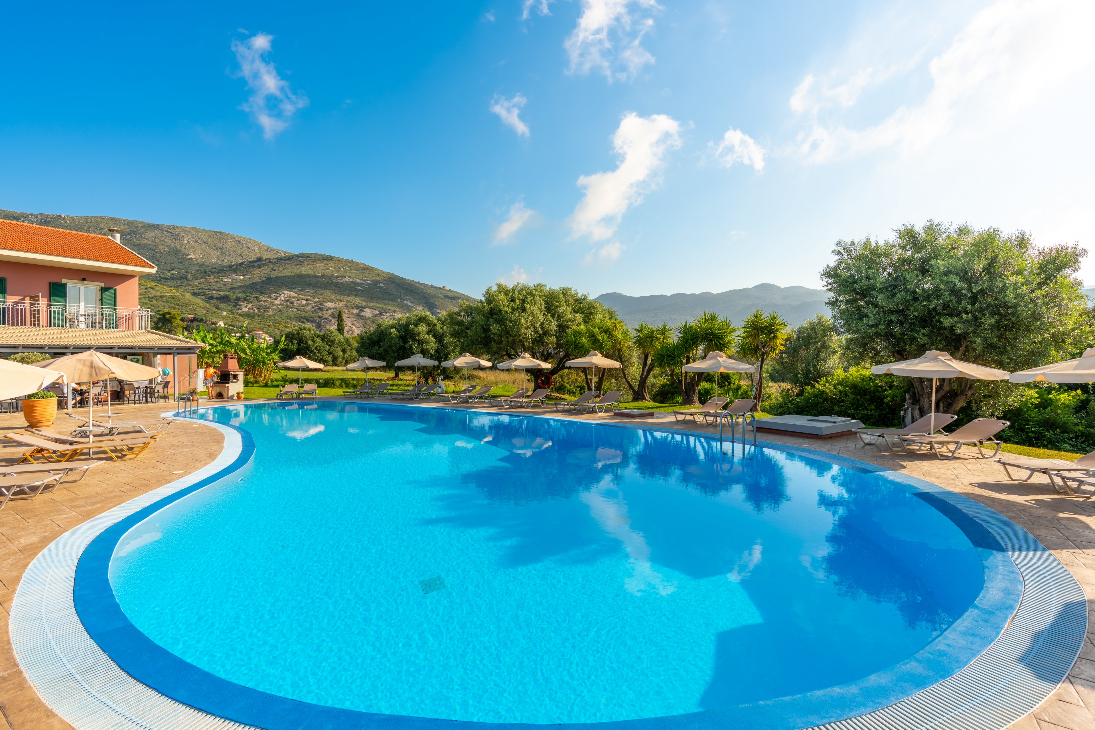 A large, clear blue swimming pool is surrounded by sun loungers and parasols in a resort setting. A warm-colored building sits to the left, while lush green trees and manicured lawns frame the pool area. Rolling hills and mountains rise in the background under a bright blue sky with scattered clouds. The scene evokes a relaxing vacation atmosphere in a sunny, tranquil location.