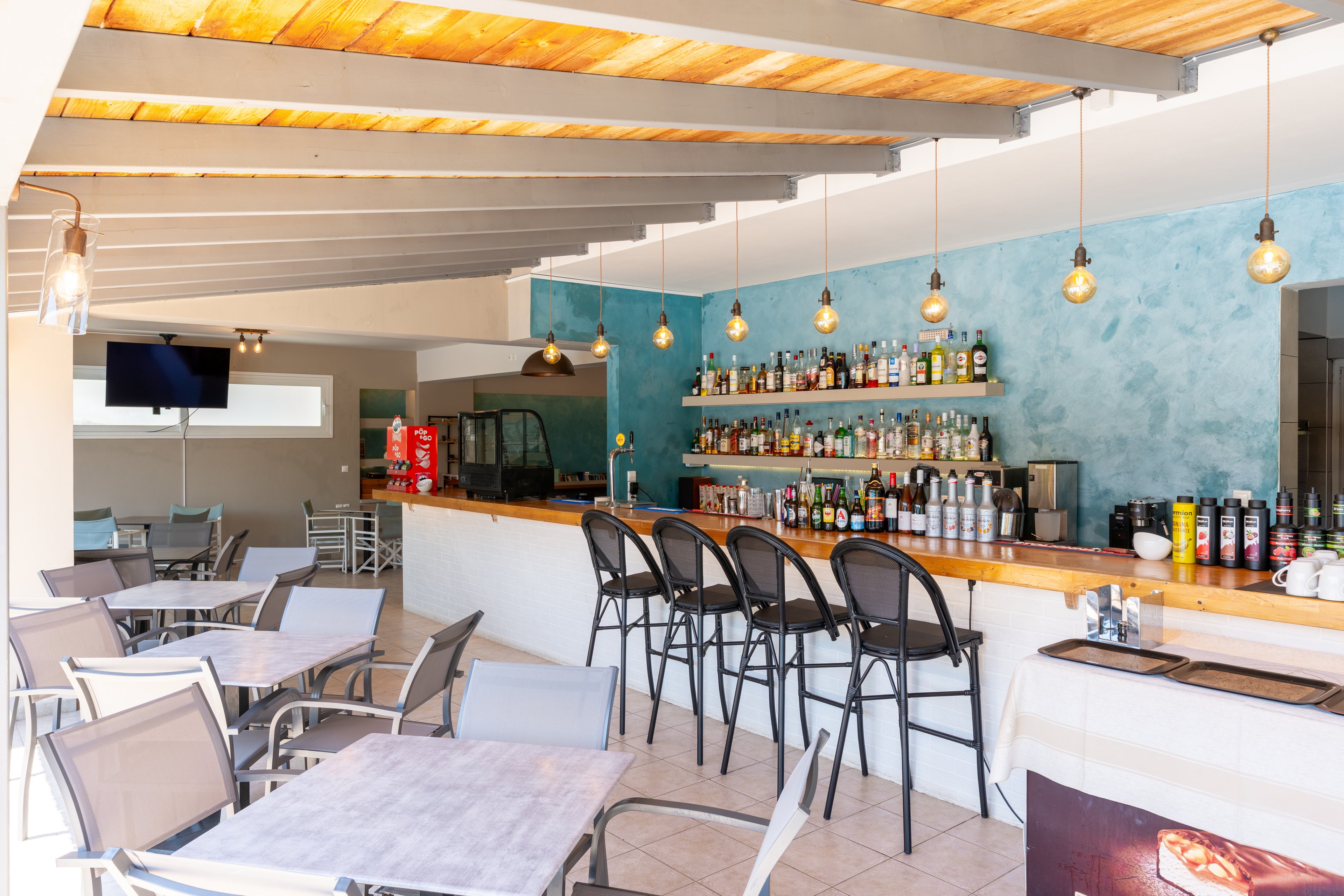 A contemporary bar interior features a long wooden counter lined with high metal stools and shelves filled with assorted bottles. Pendant lights hang from the ceiling, casting warm light over the bar and nearby tables with metal chairs. A turquoise accent wall adds color to the bright, open space, complemented by a light wood ceiling and tiled floor. In the background, a television screen and beverage cooler are visible, suggesting a casual, relaxed atmosphere.