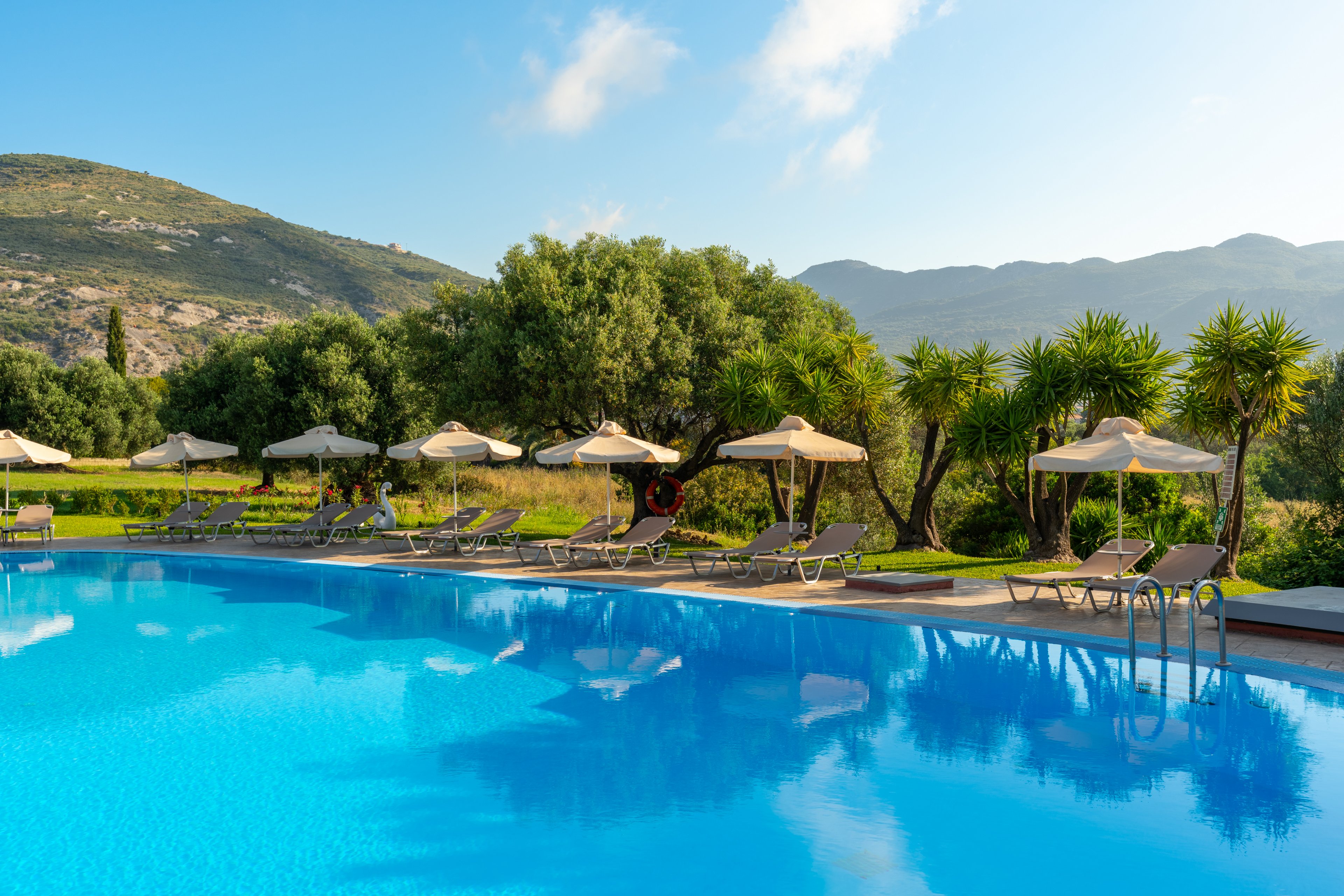 An outdoor swimming pool reflects the clear blue sky in a sunny resort setting. Lounge chairs and beige sun umbrellas line the poolside, facing lush green trees and shrubs. Rolling hills and distant mountains create a scenic natural backdrop. The tranquil atmosphere suggests a relaxing vacation spot in warm weather.